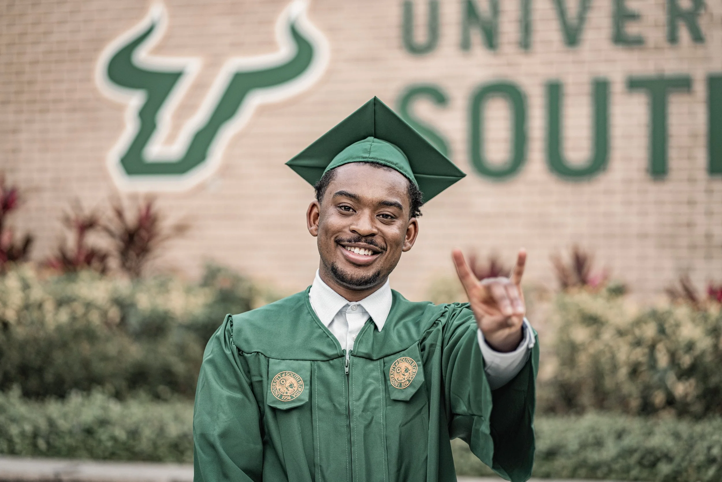 A graduate in a green cap and gown smiling and making a peace sign at graduation.