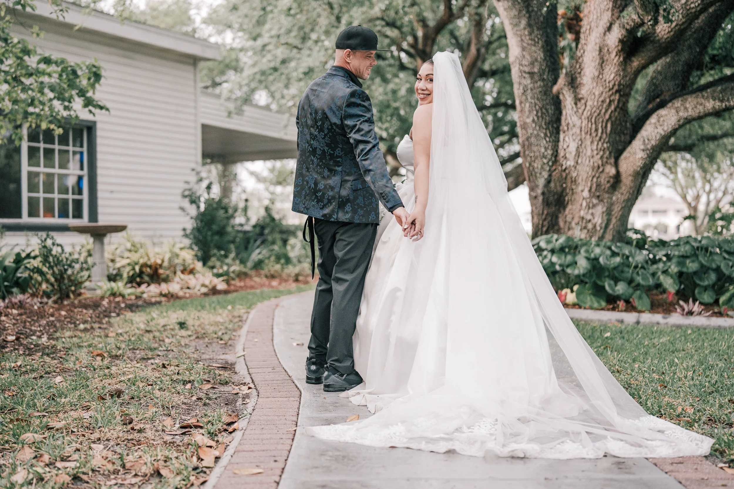 A bride and groom holding hands and smiling outdoors during a wedding. The bride is wearing a white wedding gown with a long veil, and the groom is in a dark, patterned jacket and a black cap. They are standing on a sidewalk with a large tree and gre