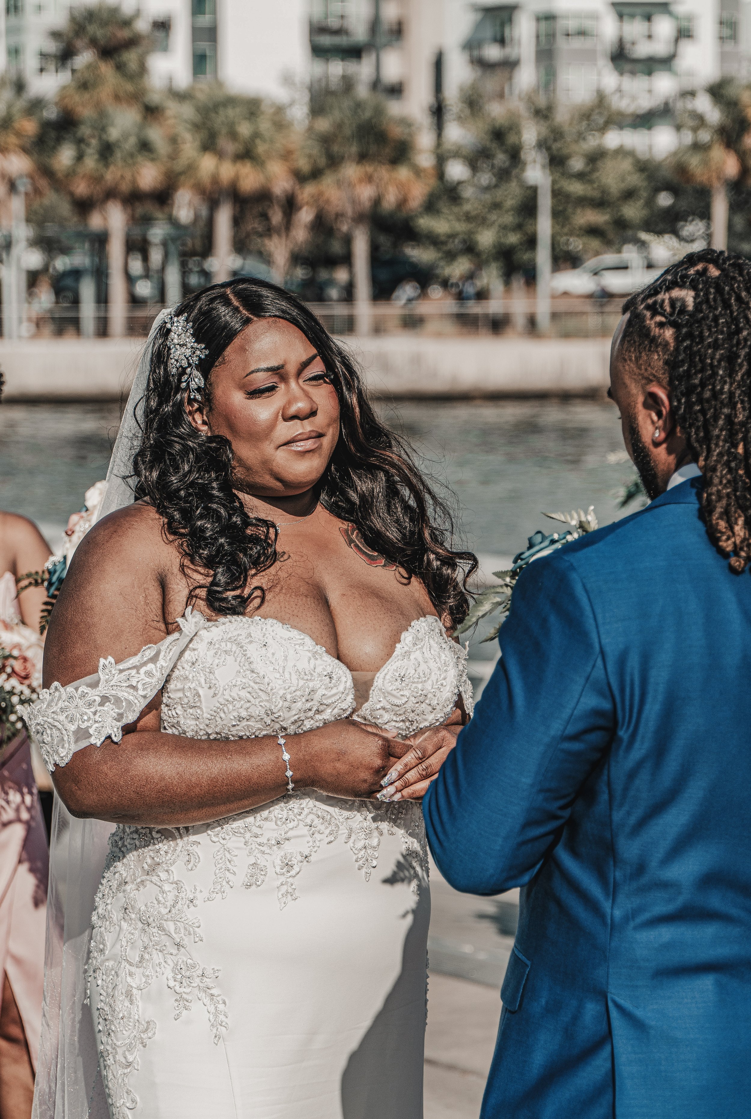 A wedding ceremony outdoors, showing a bride in a white lace wedding dress and a groom in a blue suit holding hands, with a riverside background and trees.