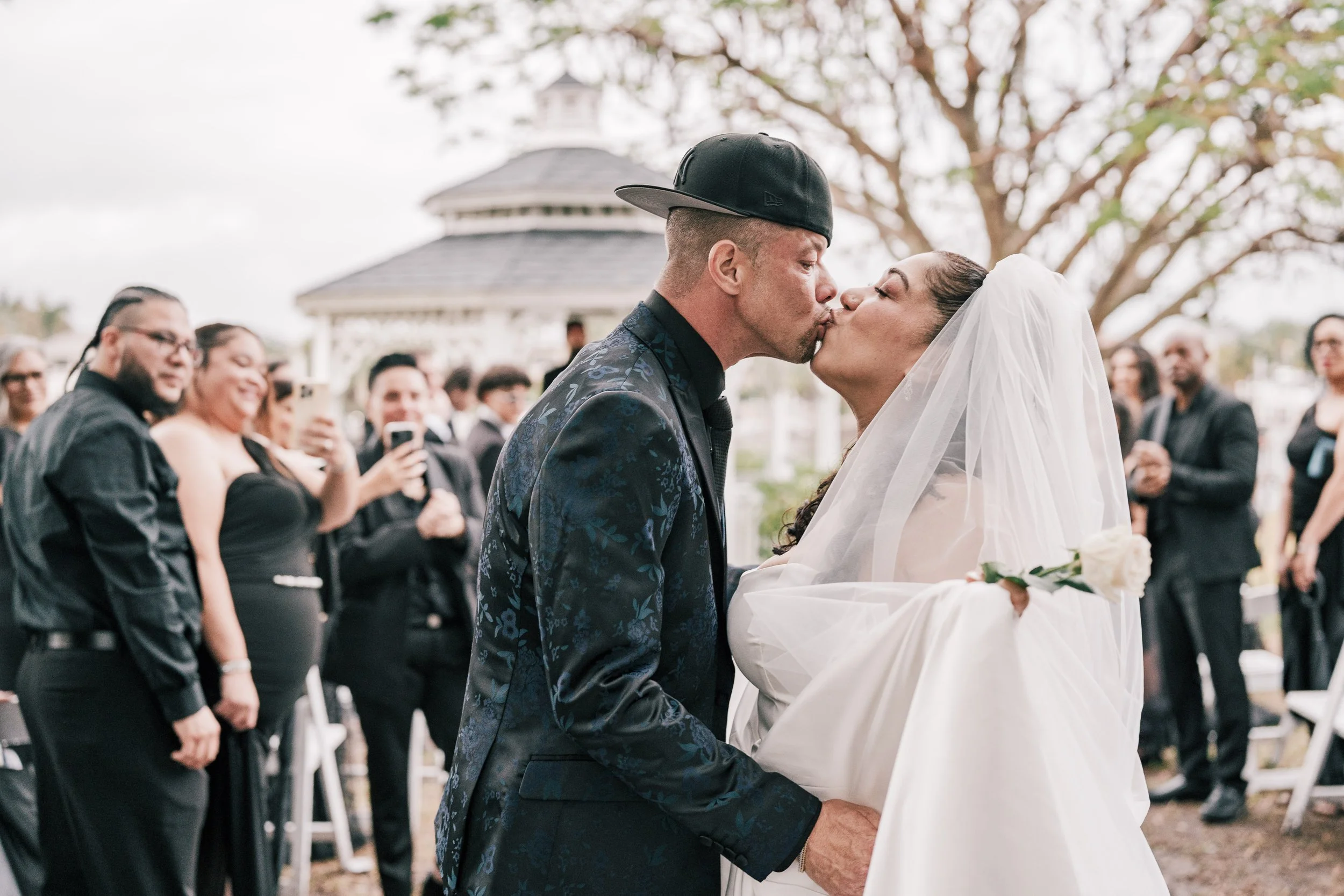 A couple sharing a wedding kiss outdoors during their wedding ceremony, with guests watching and taking photos in the background.