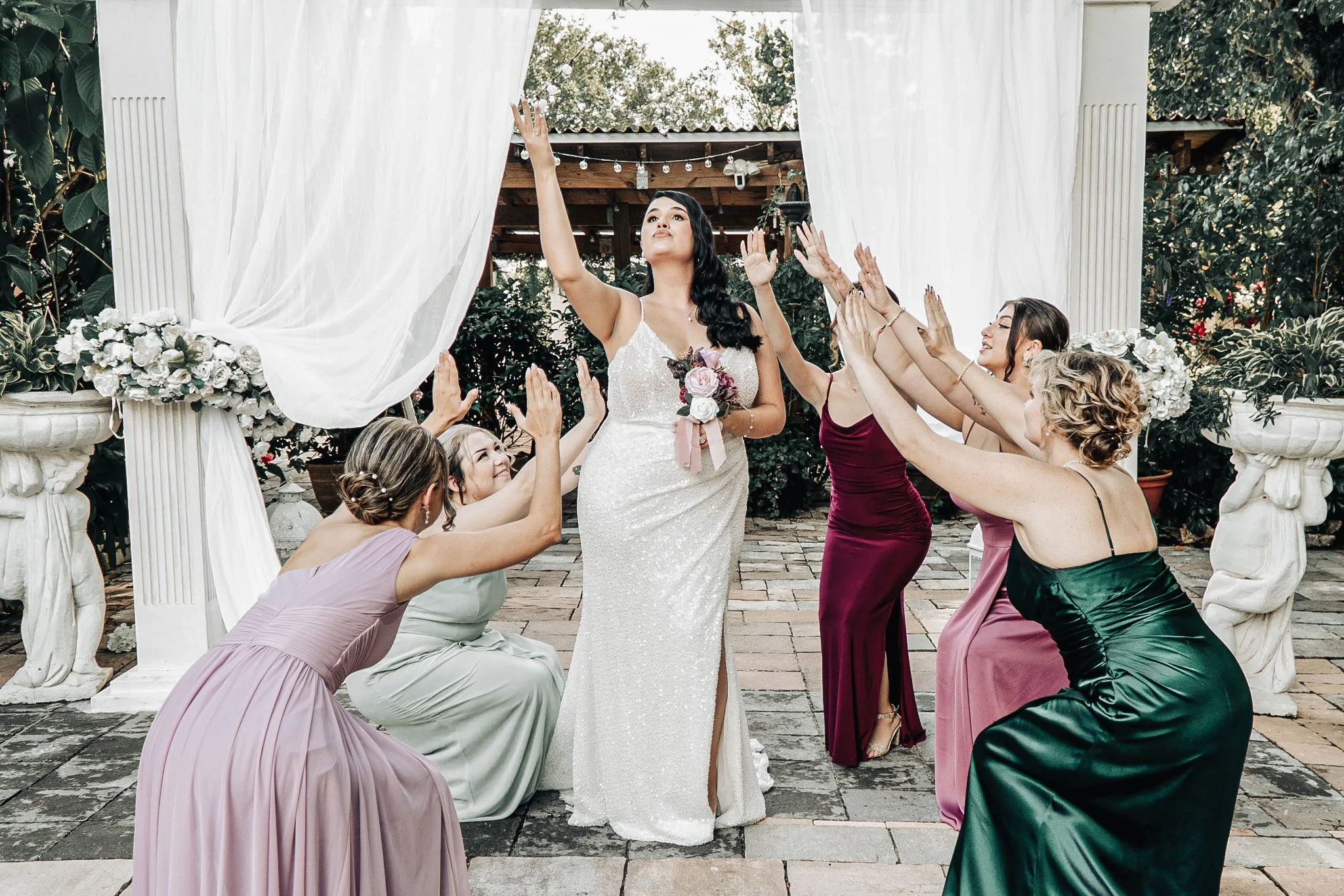 A bride in a white wedding gown with a bouquet, surrounded by bridesmaids in colorful dresses, all reaching out towards her in an outdoor garden setting with white draped curtains and lush greenery.