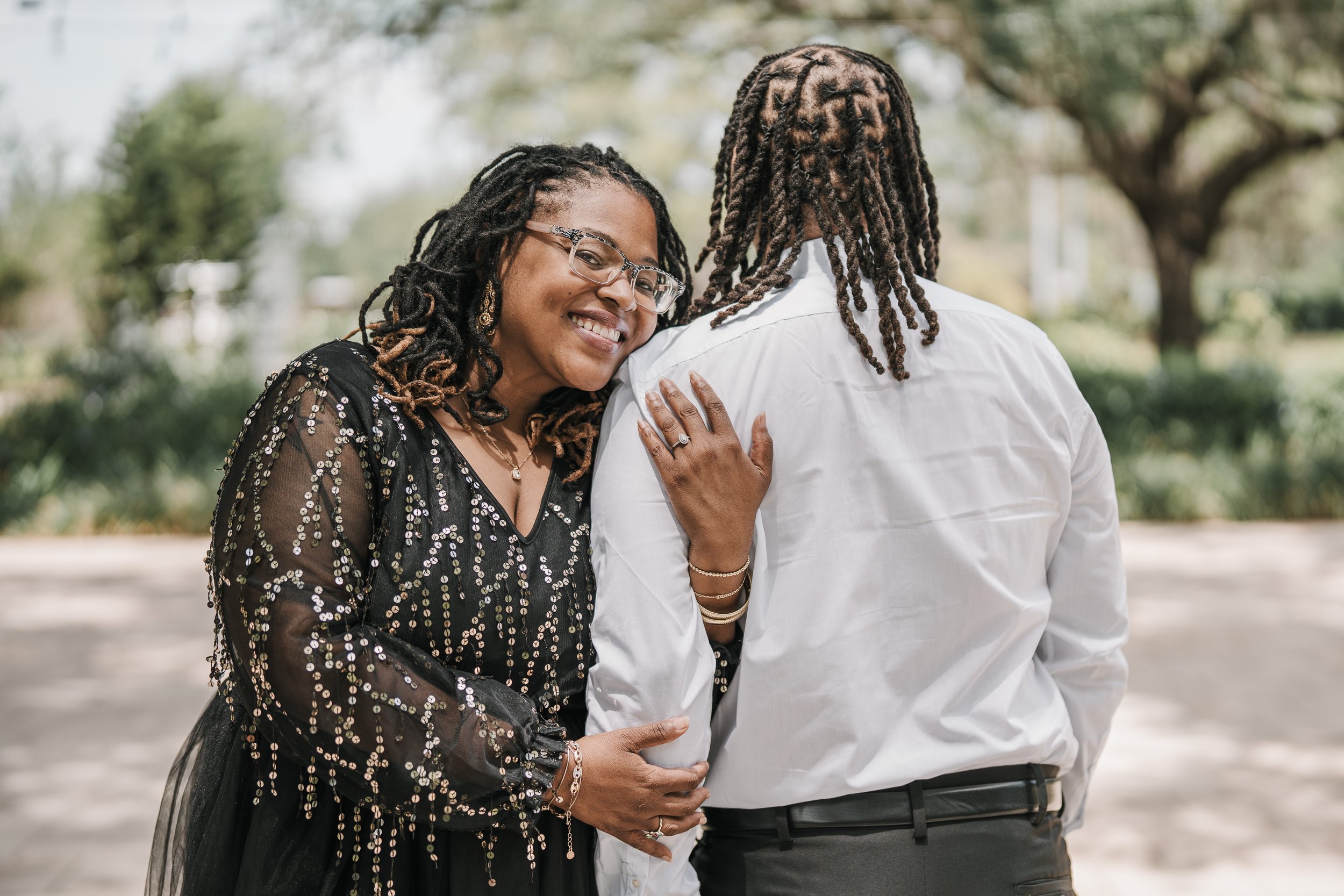 A happy woman with glasses and earrings embraces a man with long dreadlocks at an outdoor park during daytime.