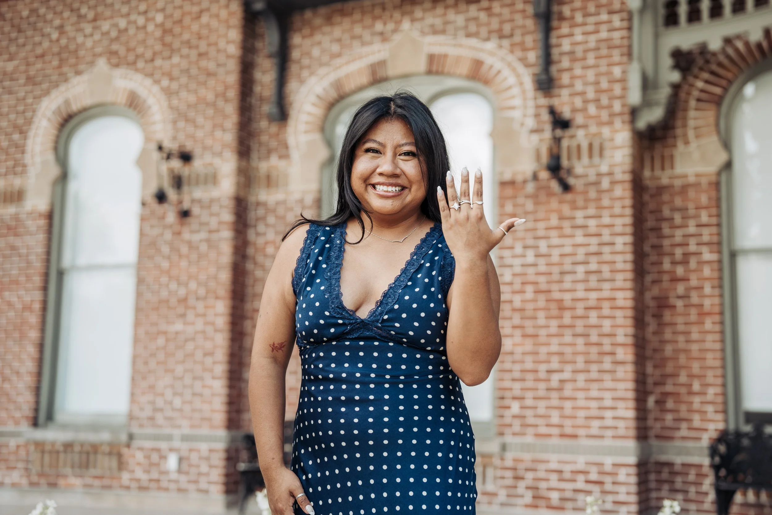 Smiling woman in polka-dot dress showing off rings on her hand outside a brick building.