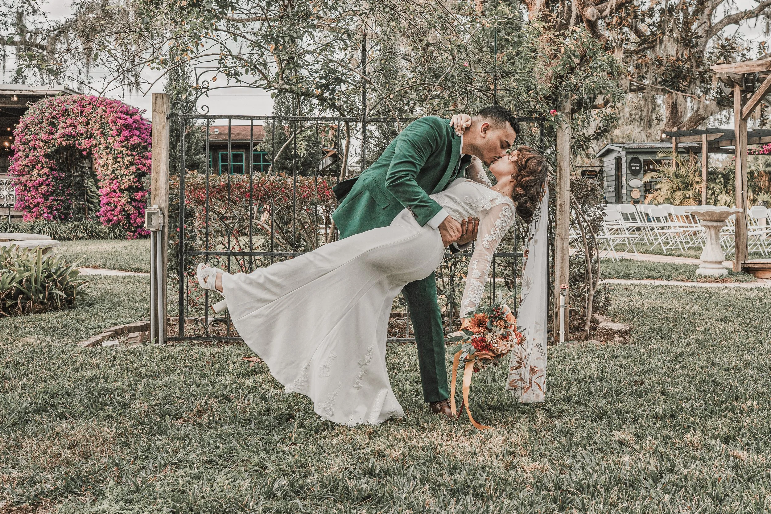 A couple sharing a kiss at their wedding outdoors, with the groom dipping the bride. The bride is holding a bouquet of flowers, and both are dressed in wedding attire. There are chairs and garden decorations in the background.