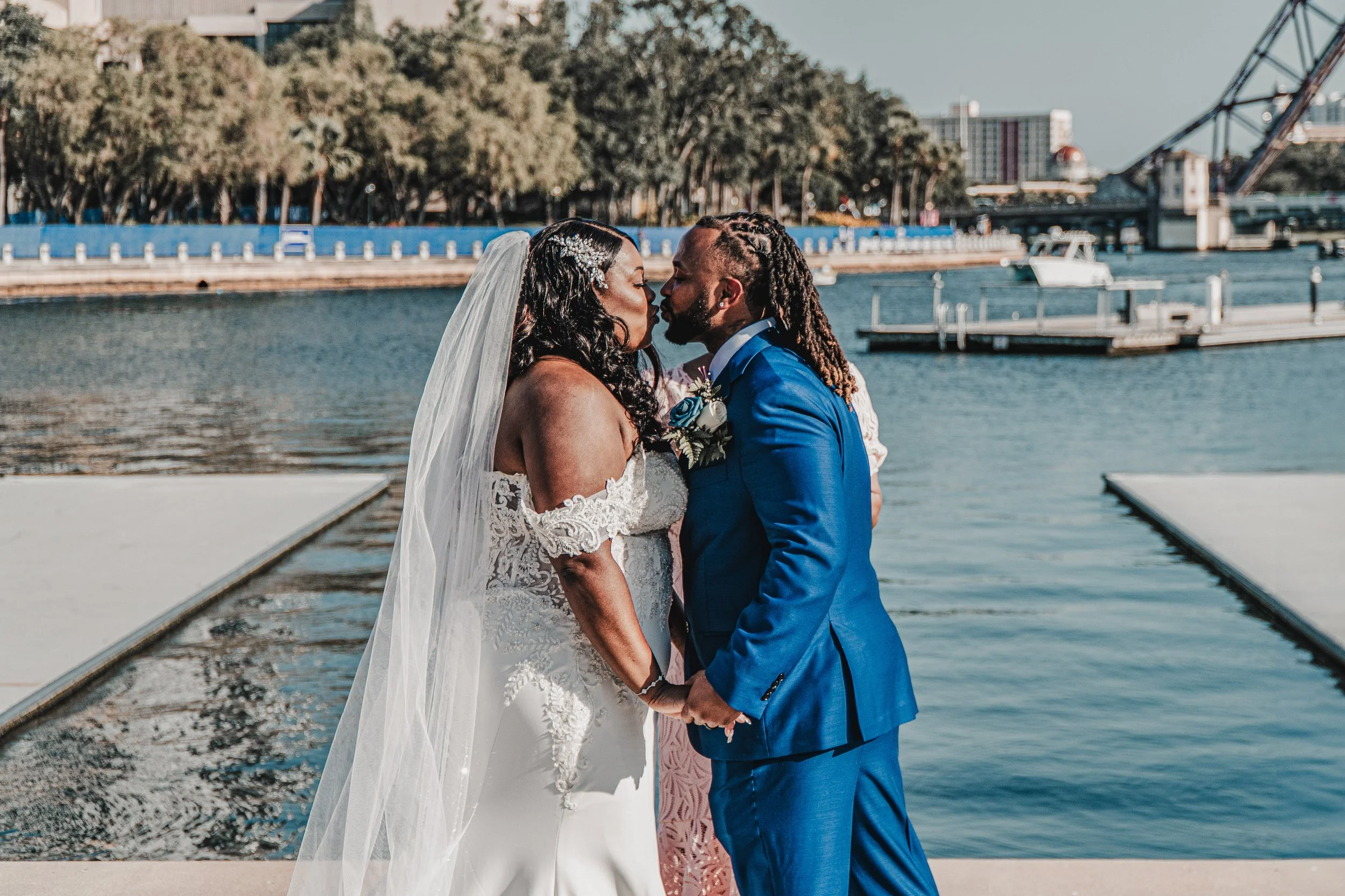 A bride and groom share a kiss by the water on a dock, with trees and city buildings in the background.