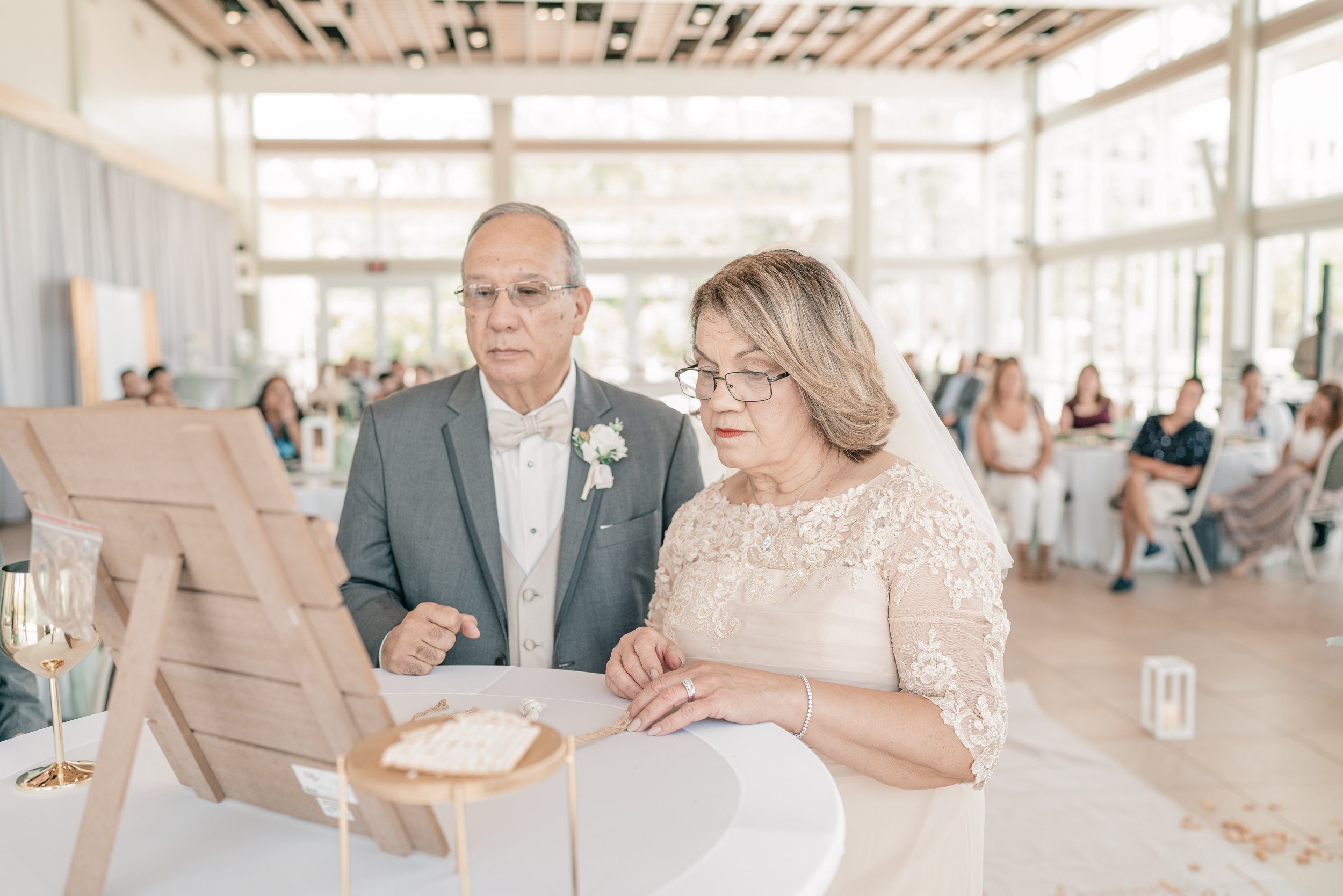 An elderly couple at a wedding, standing close together and looking at a table with wedding items, in a bright, modern venue with large windows and guests seated at tables in the background.