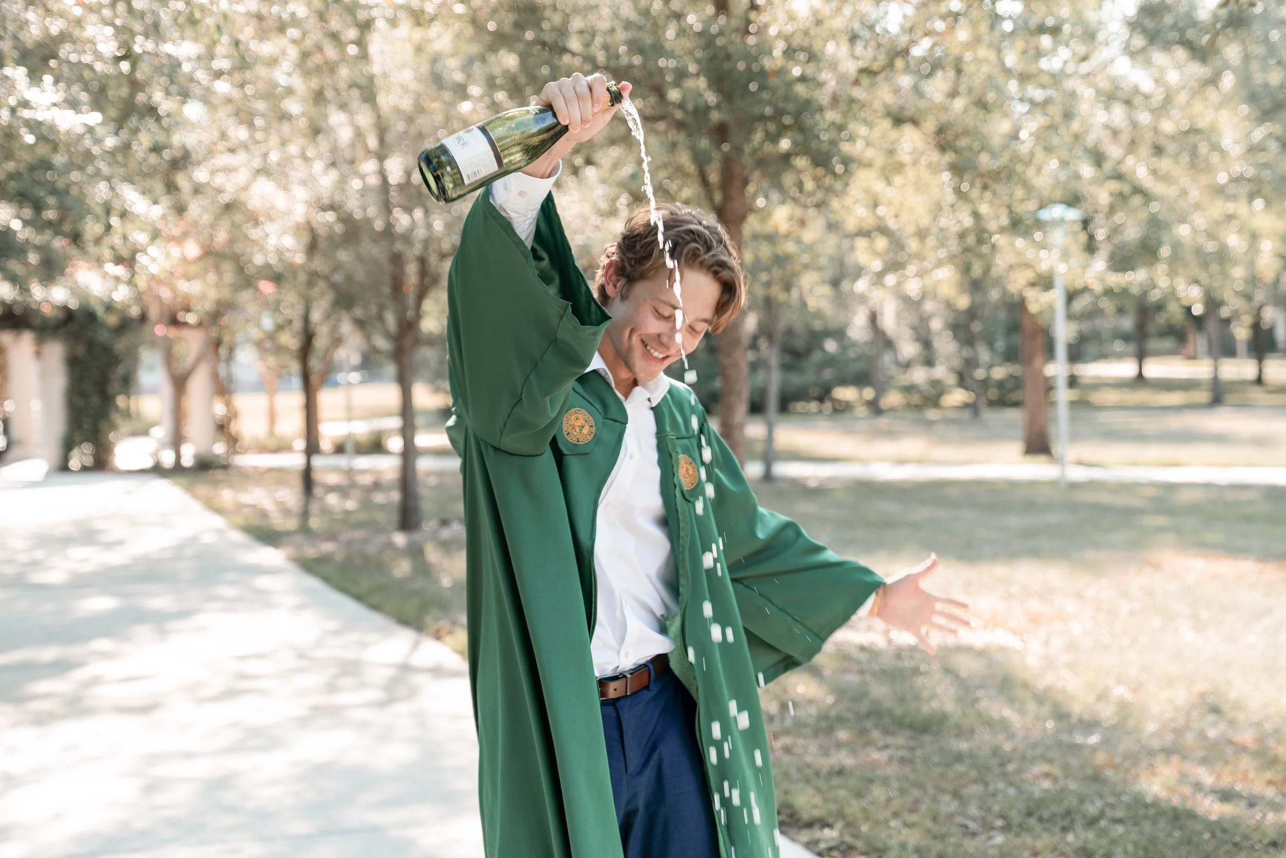 A young man in a green graduation gown smiles as he pours champagne from a bottle over his head outdoors on a sunny day, with trees and a park in the background.