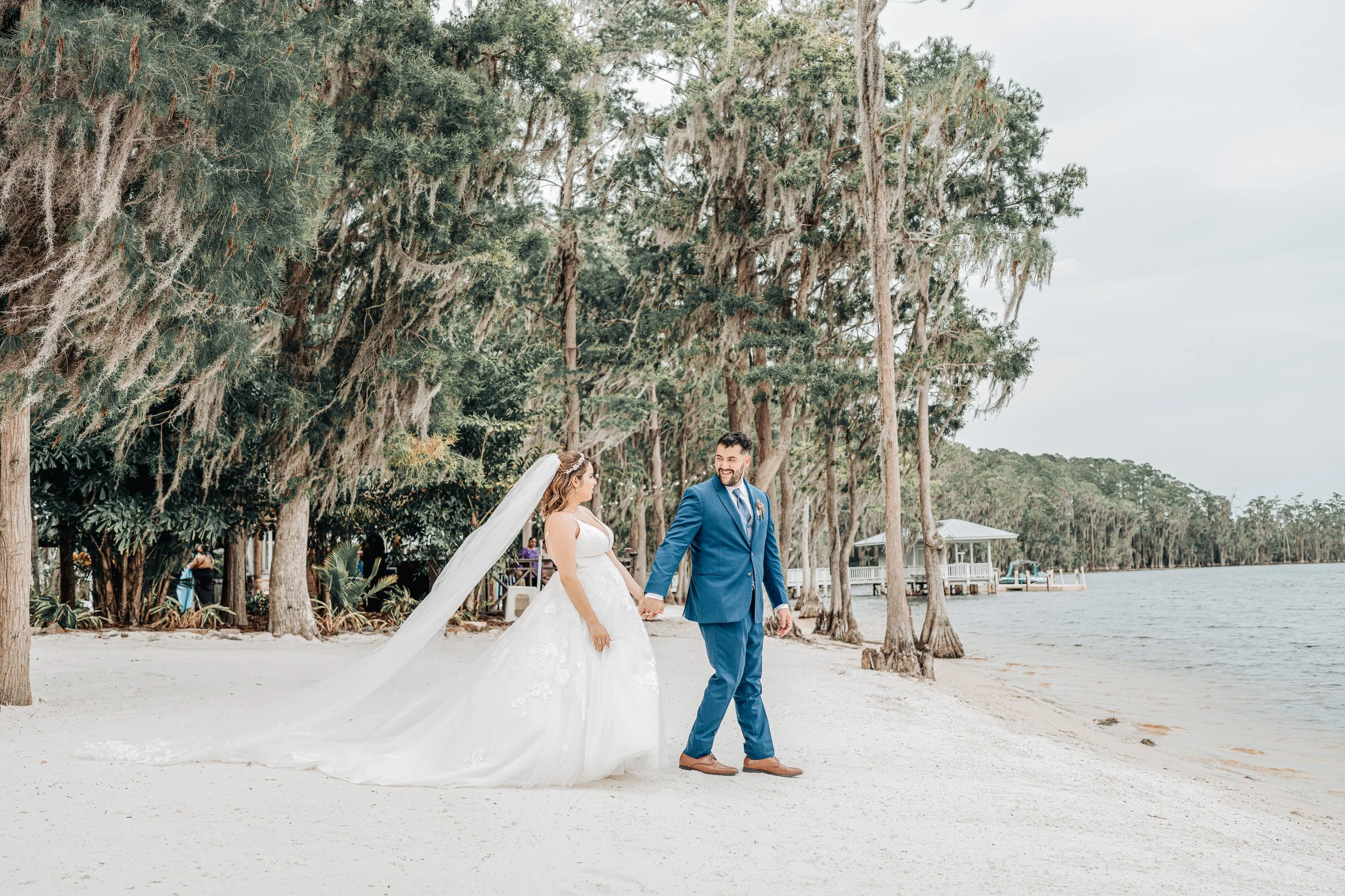 A bride and groom holding hands while walking on a sandy beach, surrounded by tall trees with Spanish moss, with water and a pavilion in the background.