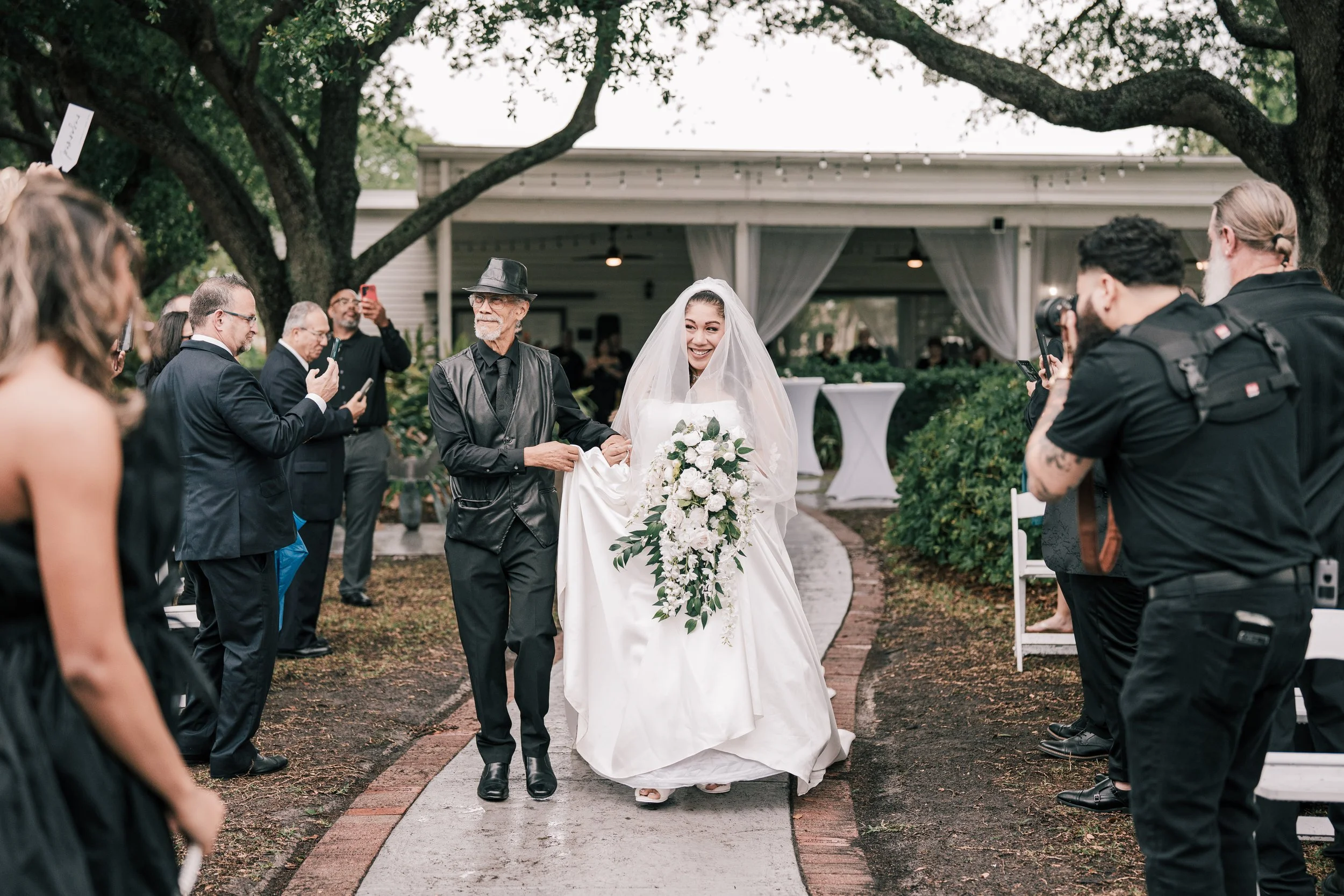 A bride walking on an outdoor pathway holding a bouquet of white flowers, accompanied by an older man, possibly her father, during her wedding ceremony, with guests on both sides capturing the moment.