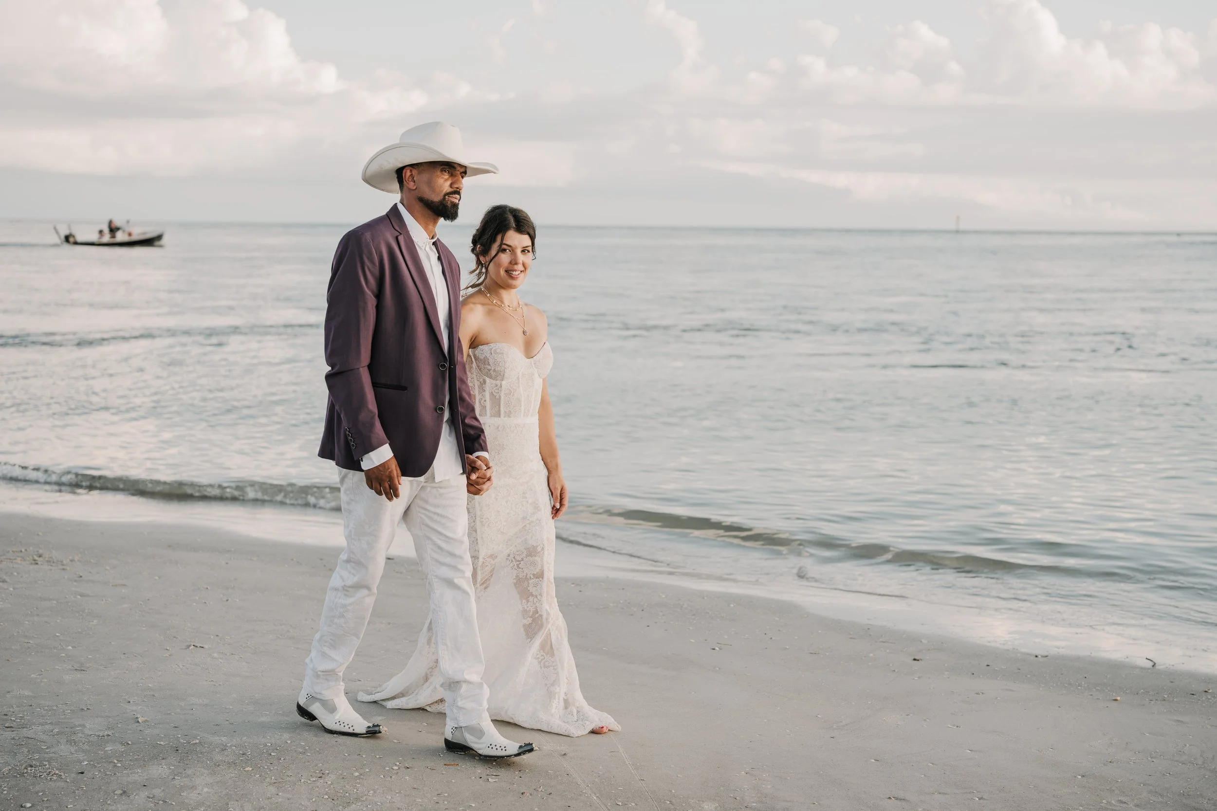 A couple standing hand in hand on the beach, with the ocean and a boat in the background. The man is wearing a purple blazer, white shirt, and white sneakers, with a white cowboy hat. The woman is dressed in a white lace wedding dress and has dark ha