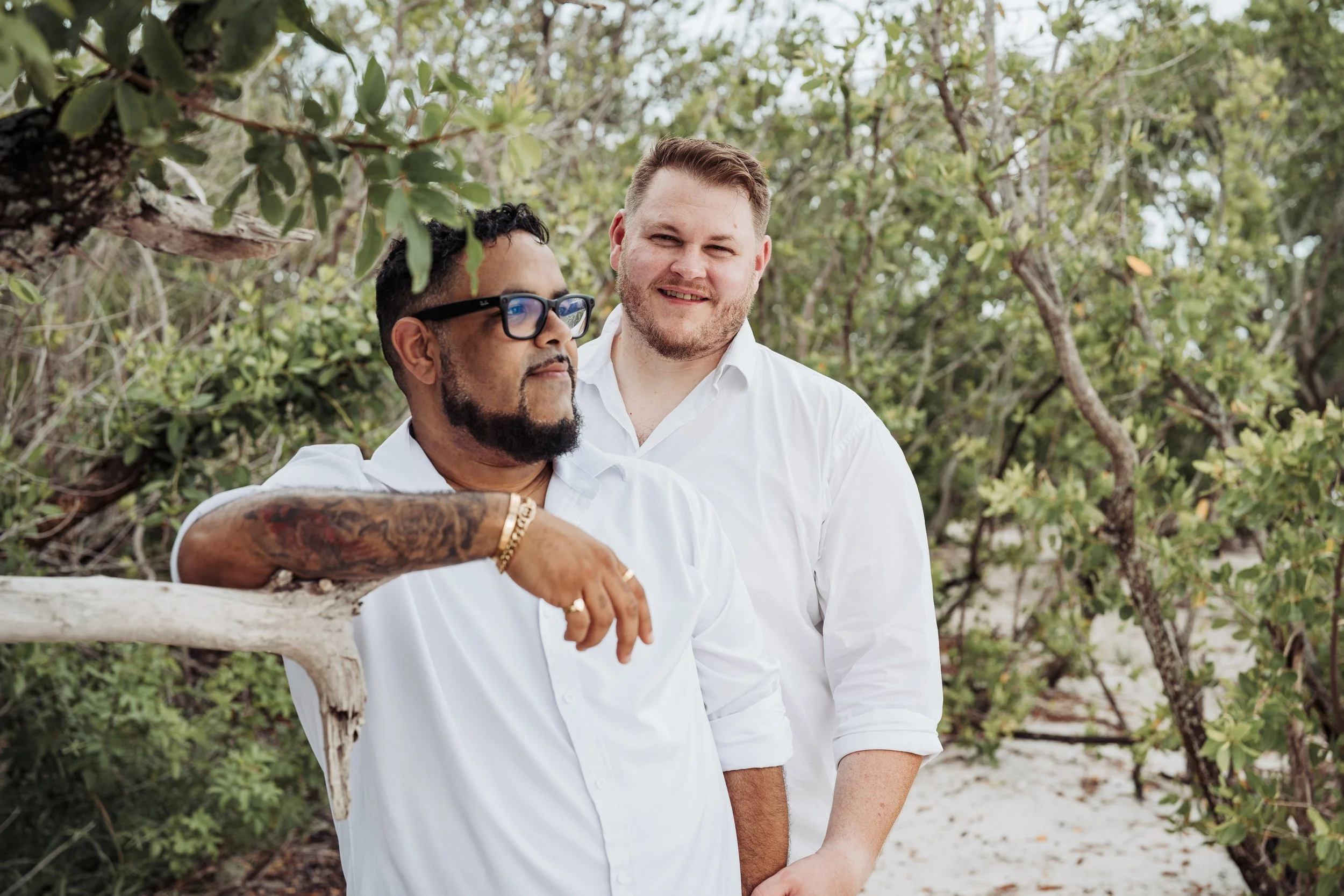 Two men in white shirts stand outdoors among trees, one with glasses and tattoos, the other smiling, with appears to be a beach or park setting.