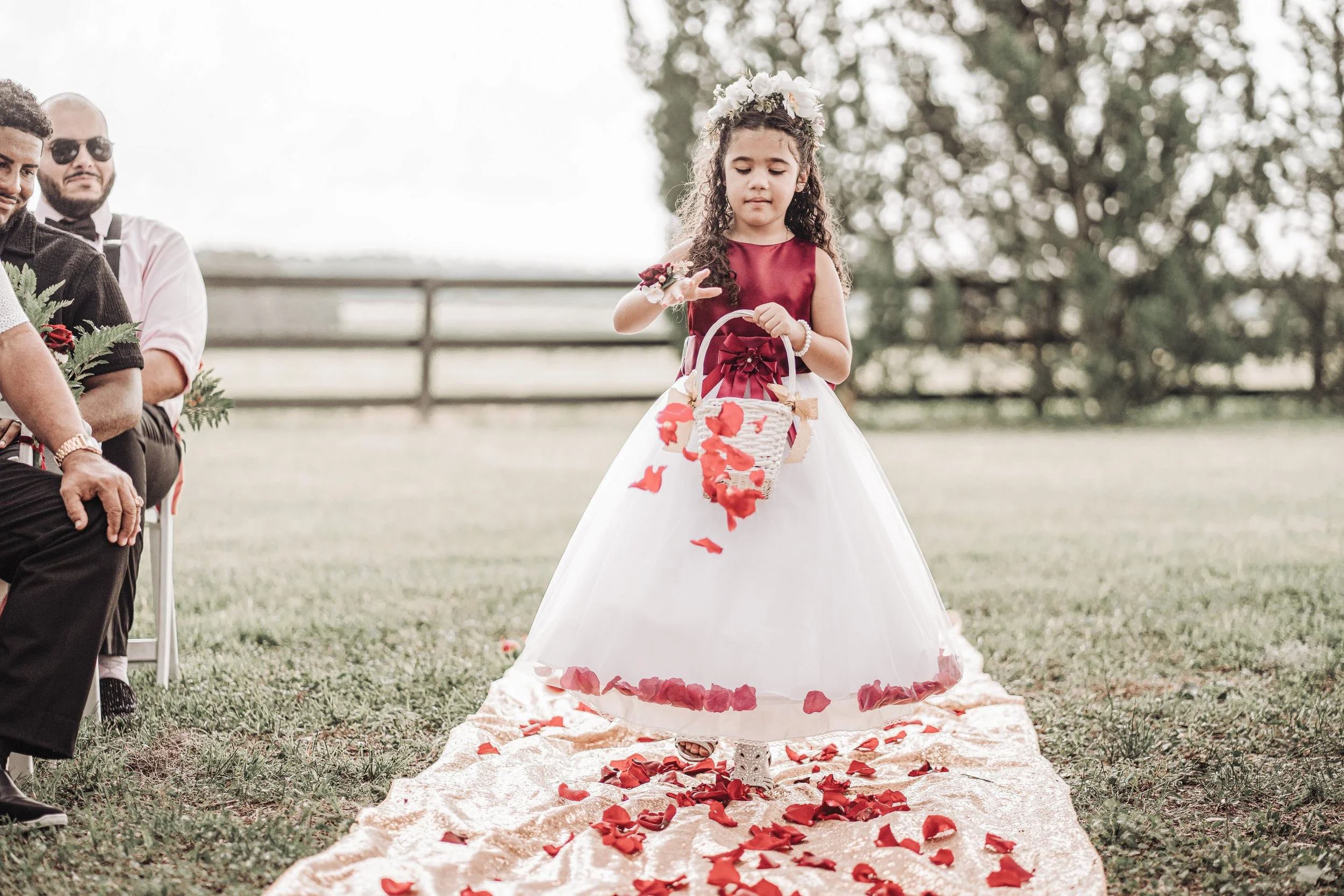 Young girl in a formal dress with a floral crown, holding a basket, walking on a rose petal-lined aisle outdoors during a wedding ceremony.