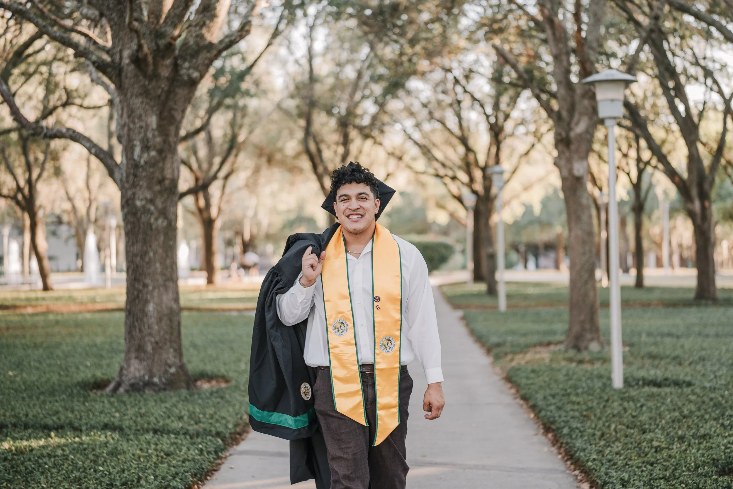 Smiling young man in graduation gown and stole outdoors on a sidewalk lined with trees.