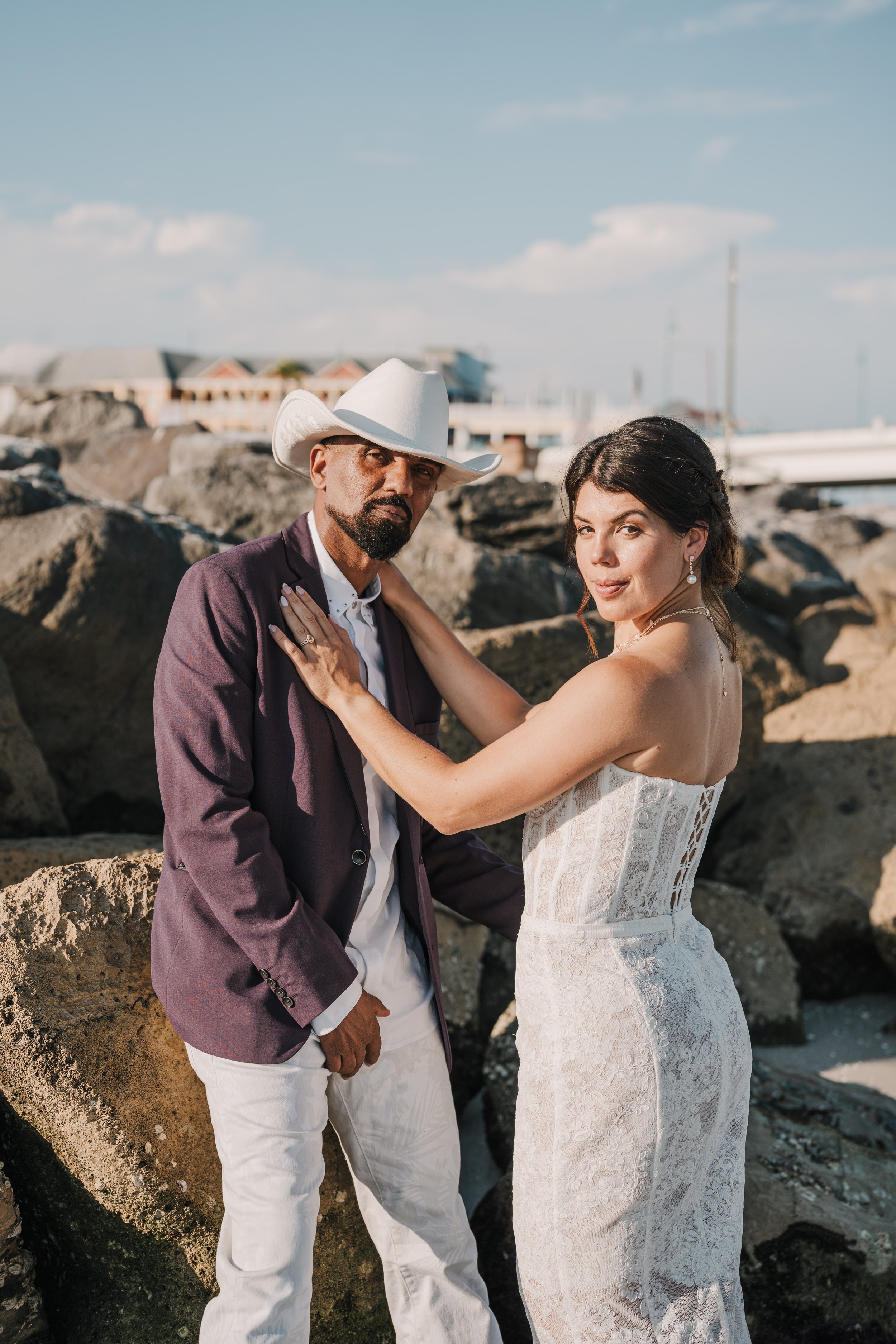 A man wearing a white cowboy hat, a purple blazer, and white pants, standing next to a woman in a strapless lace white dress, both posing on a rocky beach with buildings and a pier in the background.