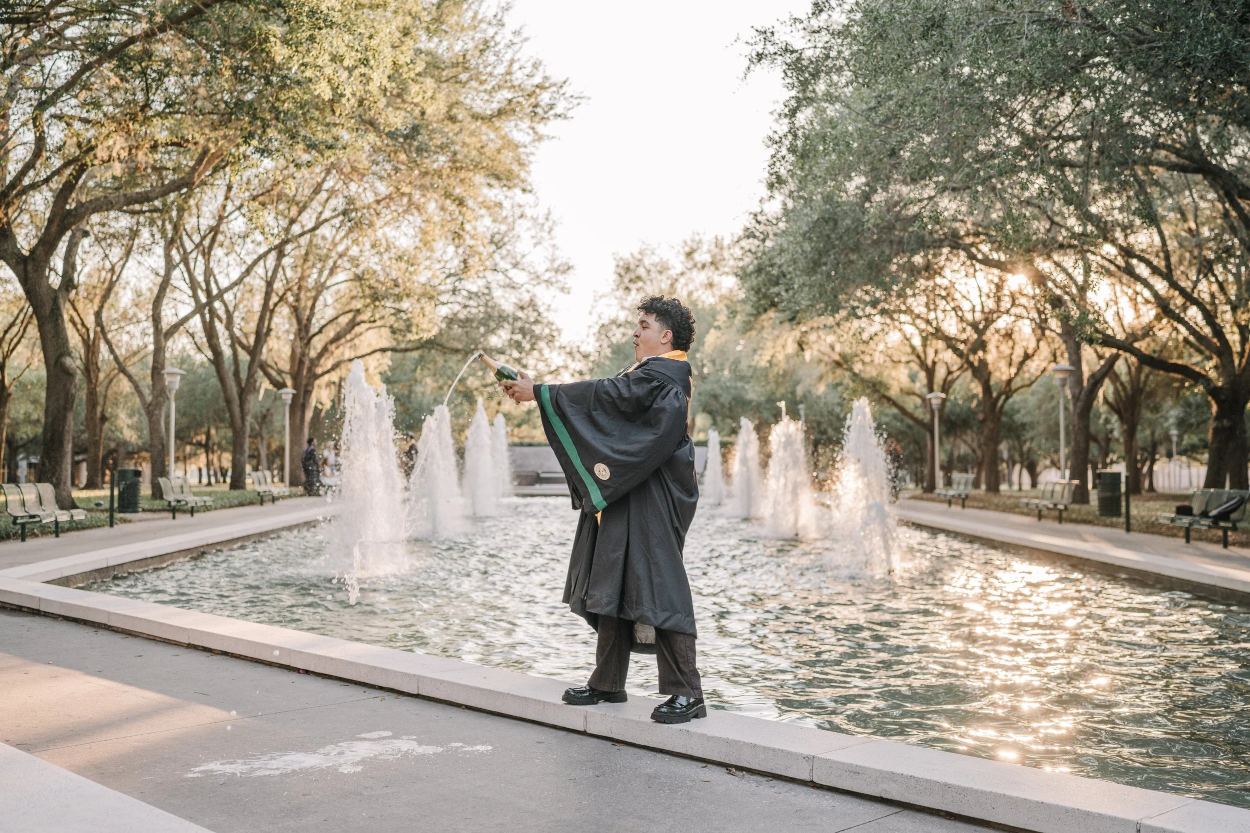 Graduate in cap and gown standing by fountain at park, pouring champagne, during sunset.