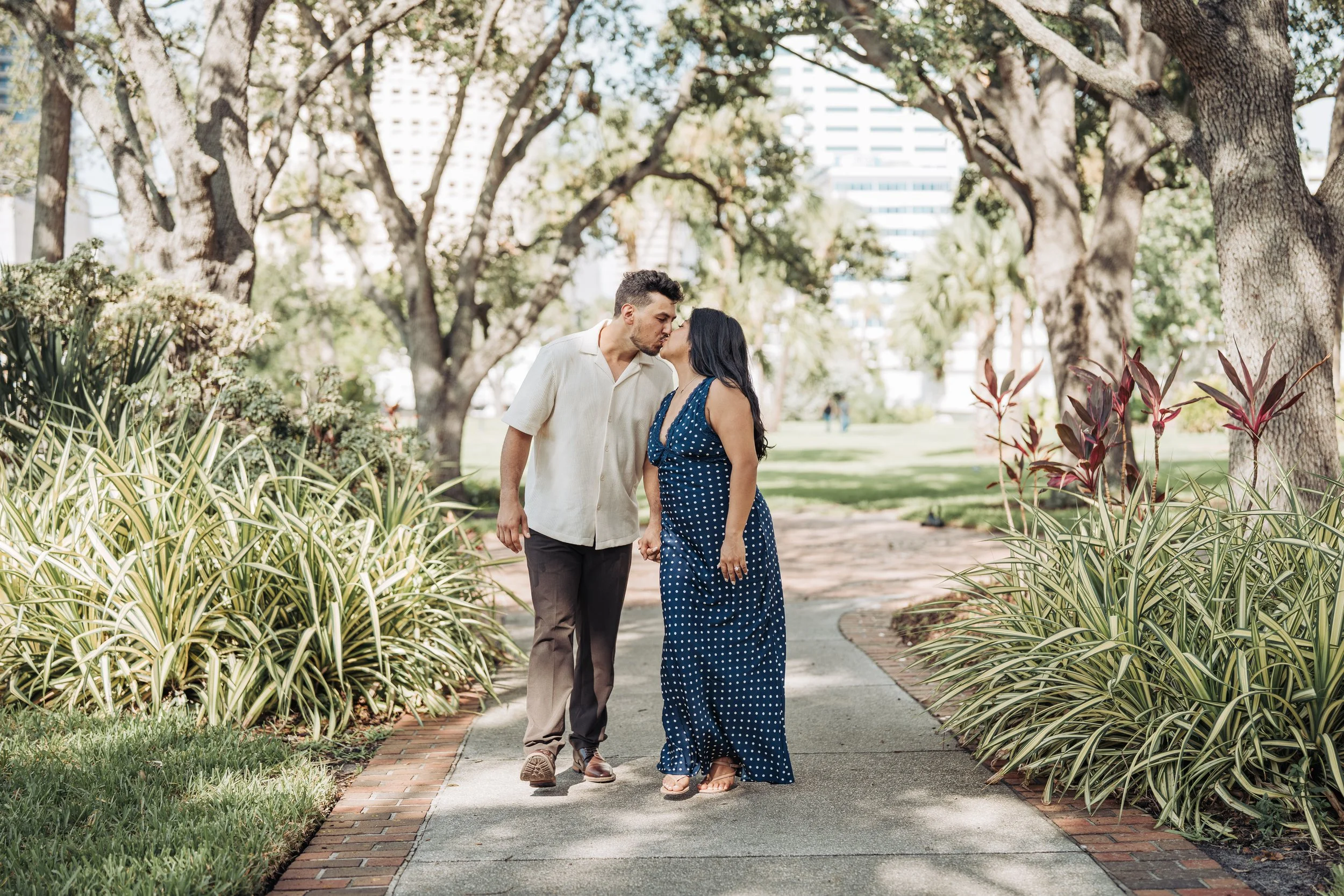 A couple holding hands and kissing while walking on a sidewalk in a park with trees and city buildings in the background.