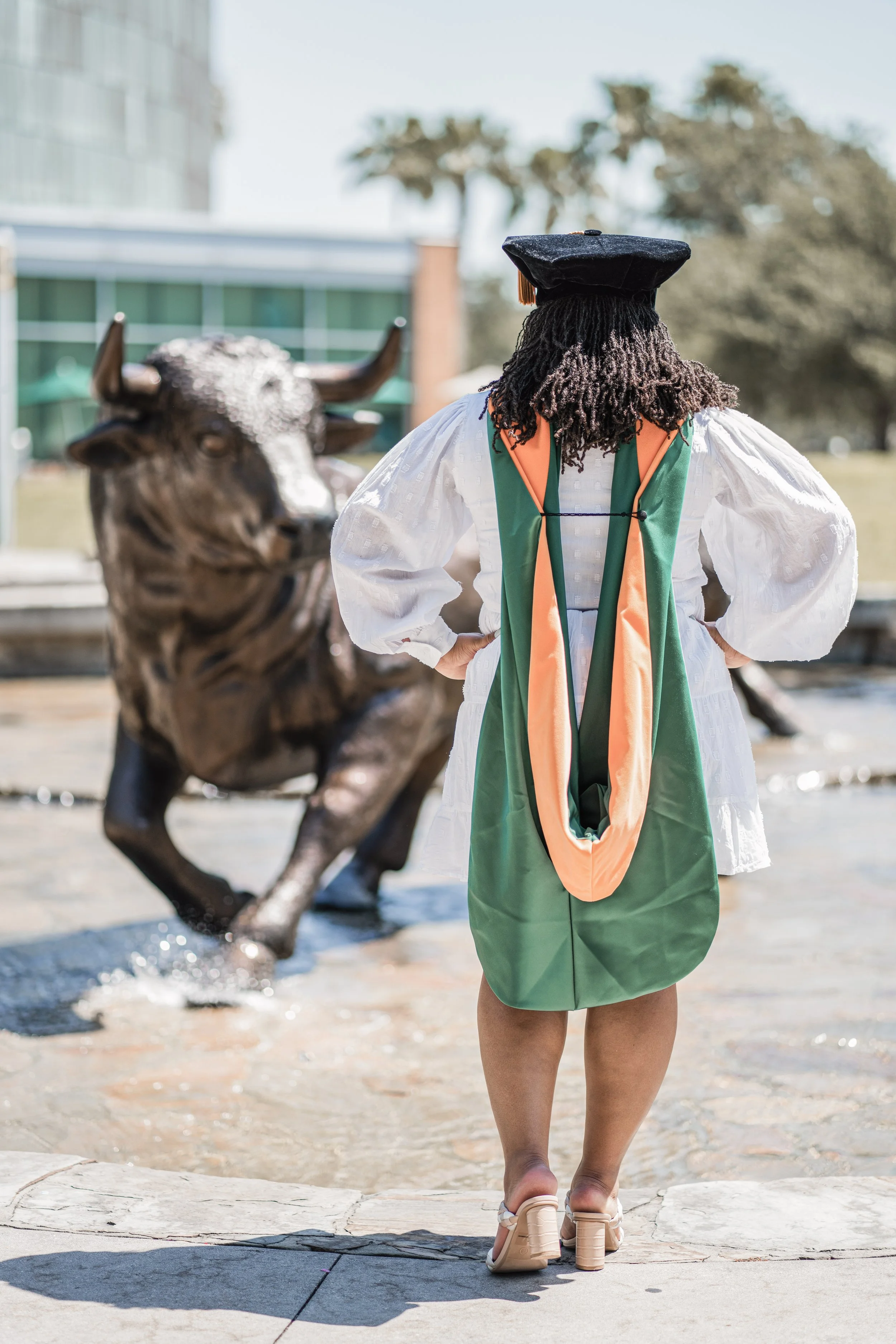 A person with curly hair wearing a graduation cap, a white dress with puffed sleeves, and a green and orange academic stole, standing by a fountain with a bronze bull sculpture. The person is facing away from the camera, with hands on hips and wearin