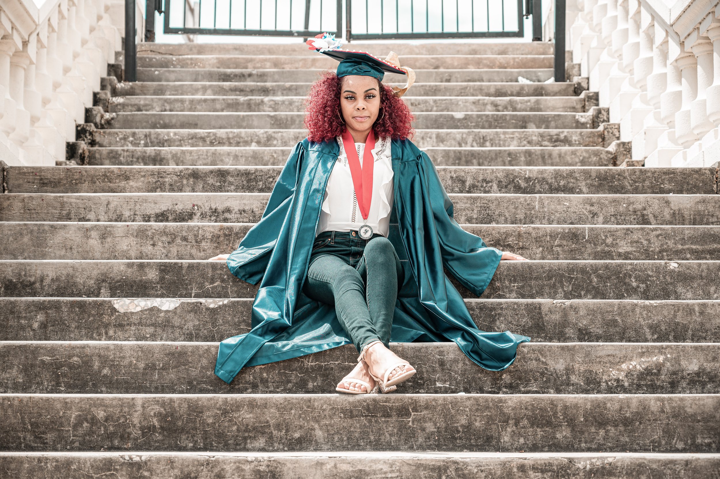 A young woman sitting on outdoor stone stairs, wearing a graduation cap and gown, with a red medal around her neck, and posing with confidence.