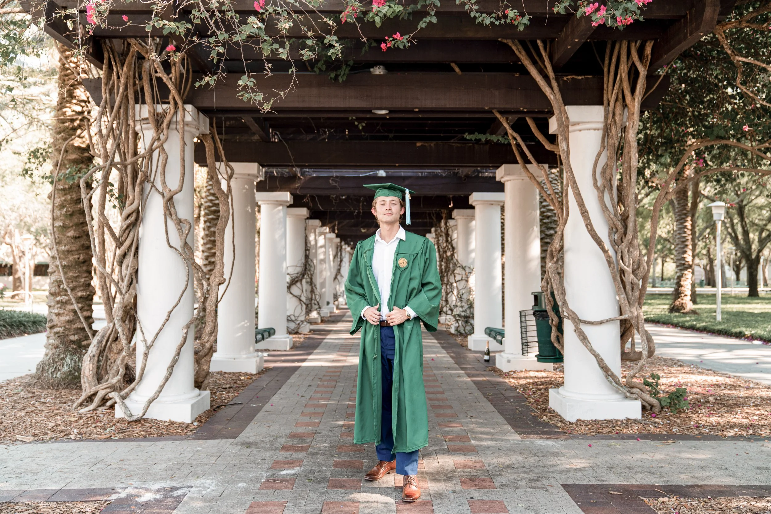 A young man in a green graduation gown and cap standing on a brick pathway under a wooden pergola with white columns and twisting vines, with pink flowers and trees in the background.