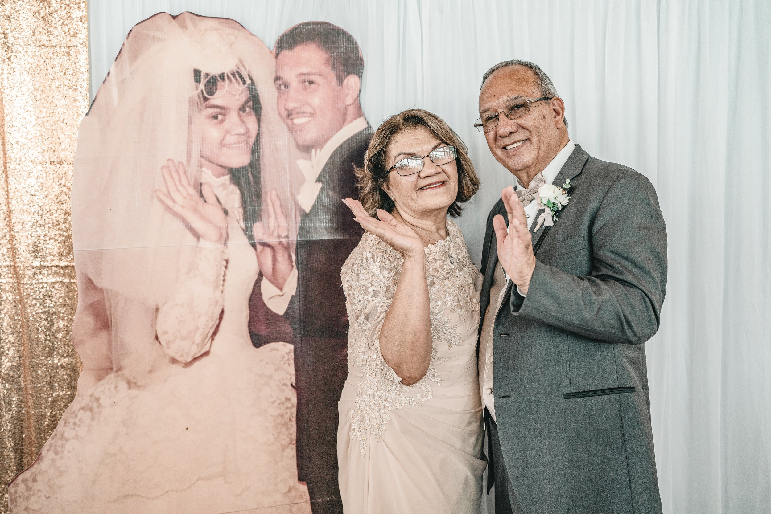 An elderly woman in a cream lace dress and an elderly man in a gray tuxedo with a boutonniere pose with a large cardboard cutout of a wedding photo featuring a bride in a veil and wedding dress and a groom in a tuxedo. The background consists of whit