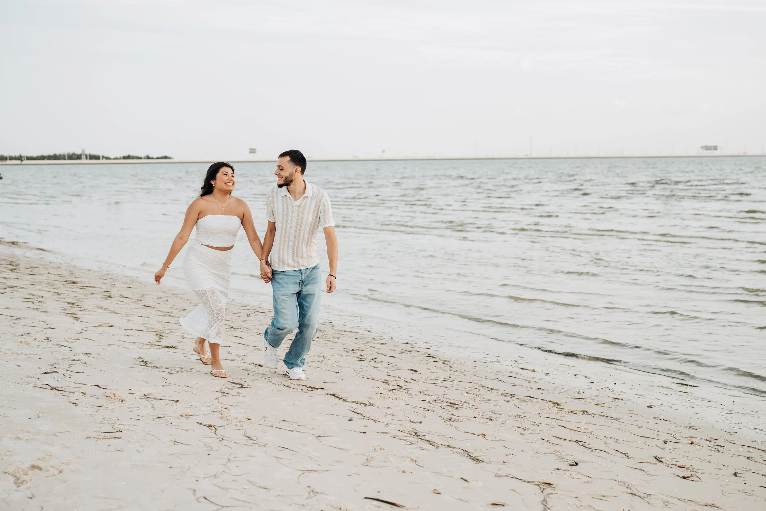 A couple holding hands and walking along a beach shoreline, smiling and enjoying each other's company on a cloudy day.