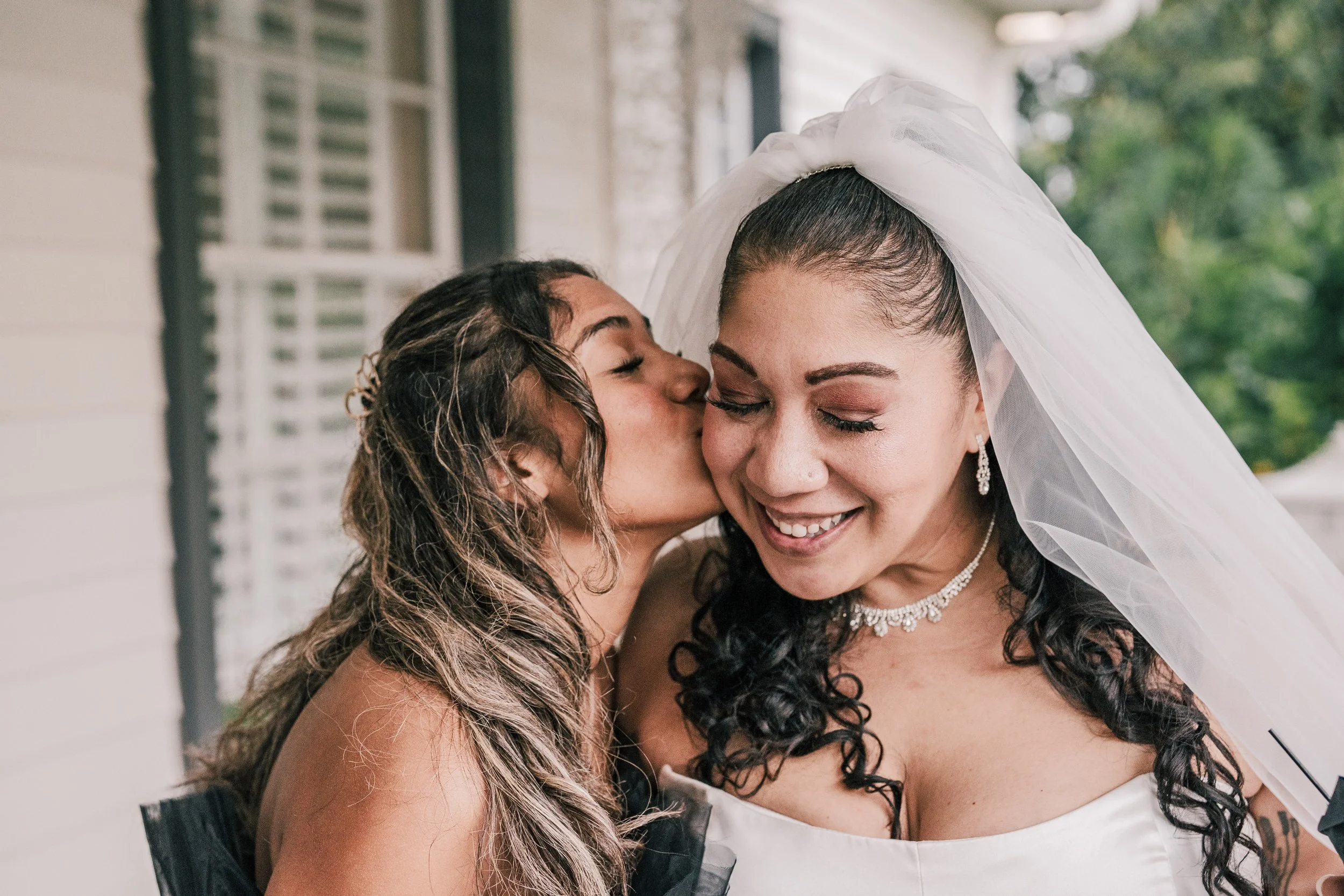 Two women sharing a kiss, one wearing a wedding veil and dress, smiling with eyes closed.