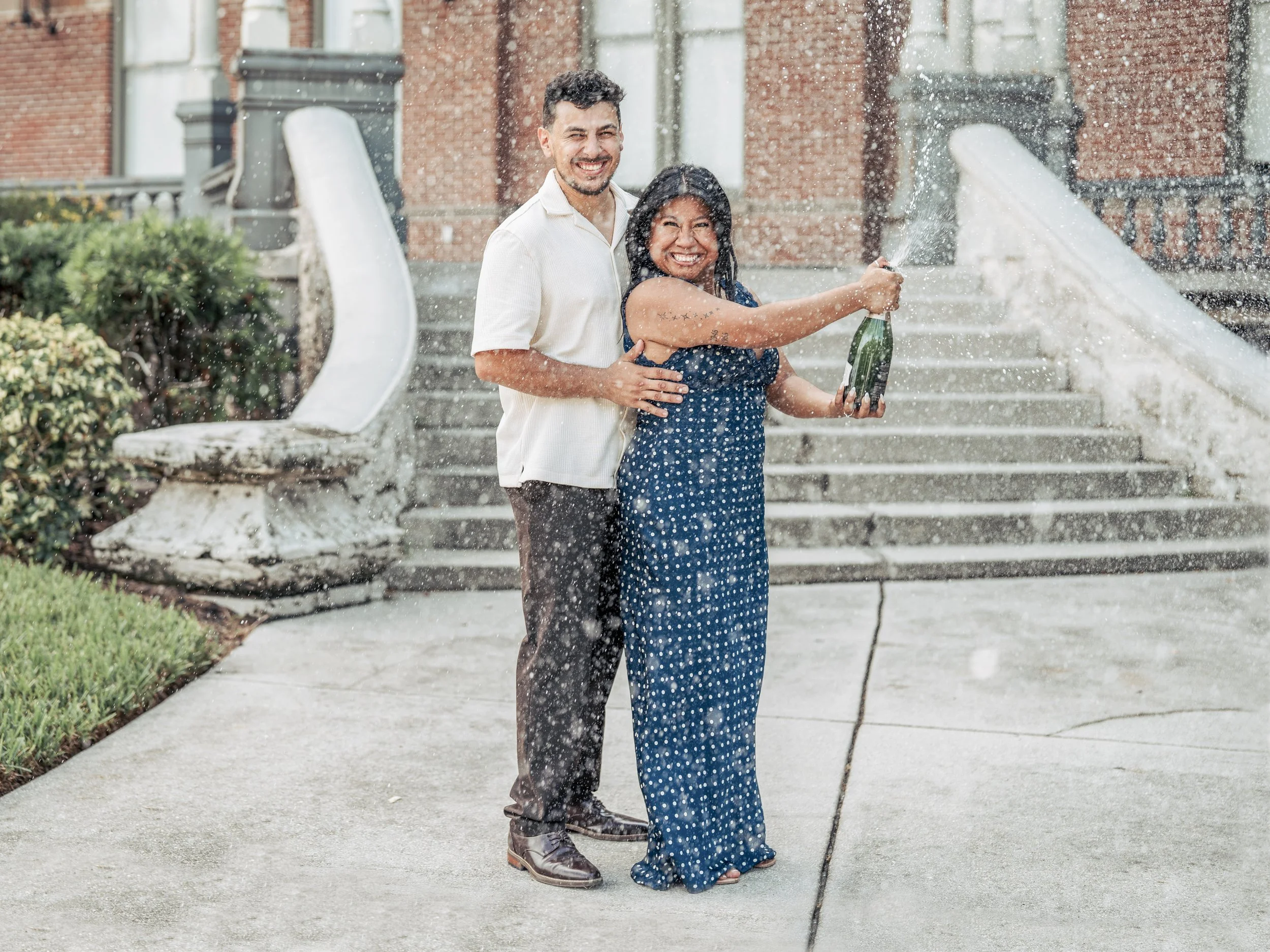 Two people celebrating on a staircase with a fountain. A man with dark hair, wearing a white shirt, hugging a woman in a blue dress with white polka dots while she holds a bottle of champagne and sprays it in the air. Both are smiling and appear happ