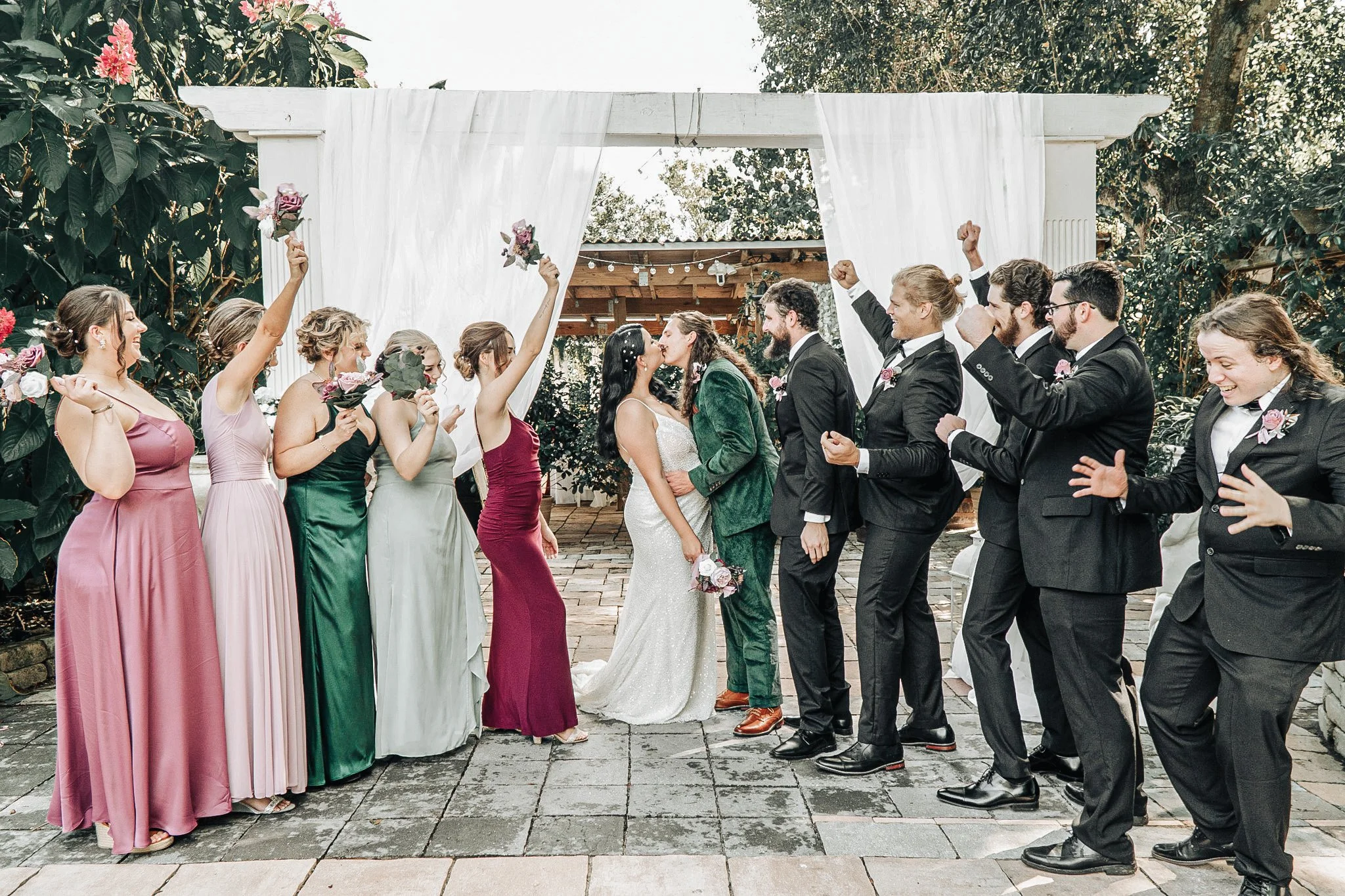 A wedding celebration outdoors with a bride and groom kissing under a white draped arch, surrounded by wedding party members holding flowers and cheering.