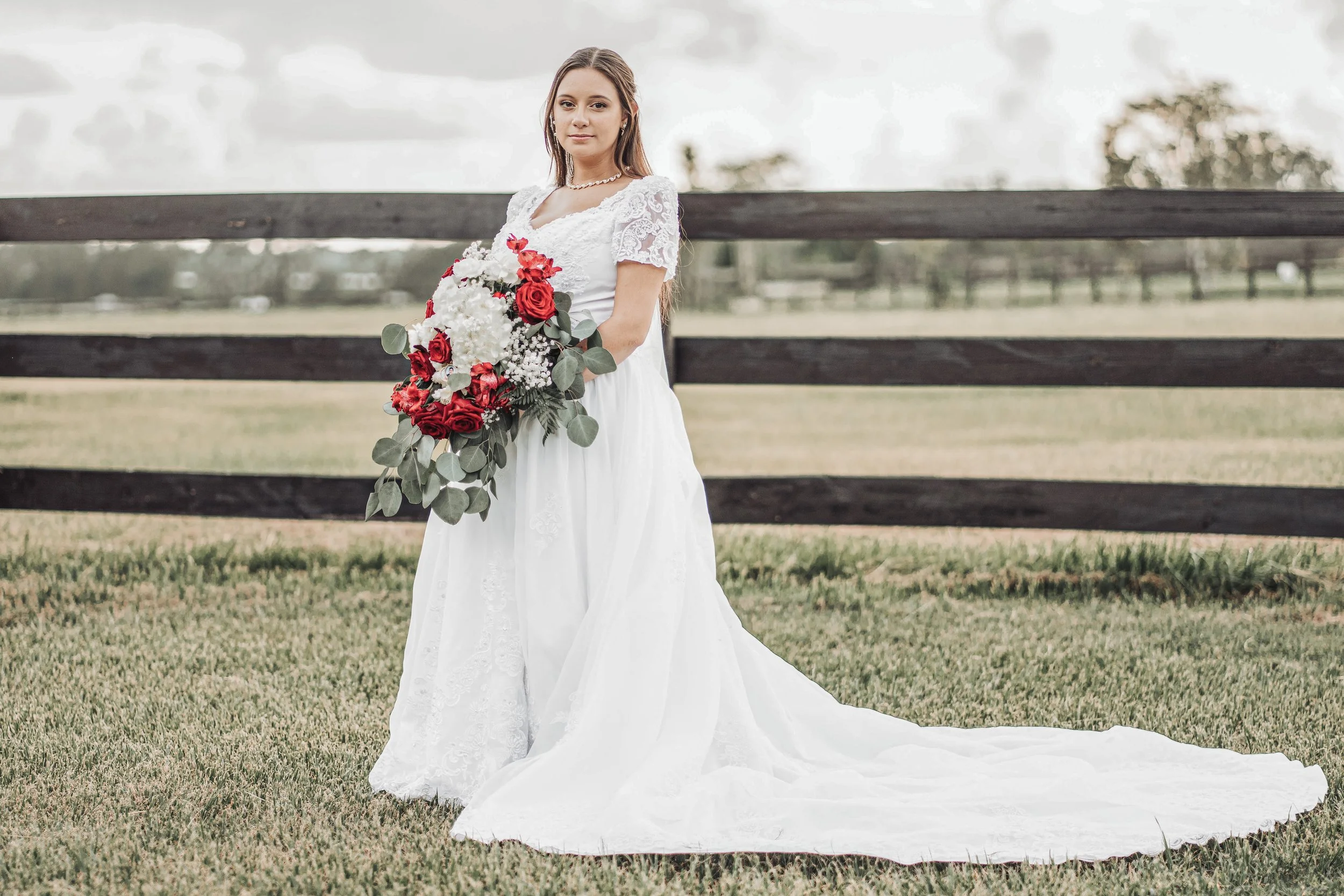 A young woman in a white lace wedding dress holding a bouquet of red, white, and green flowers stands outdoors on a grassy field near a dark wooden fence, with an overcast sky and trees in the background.