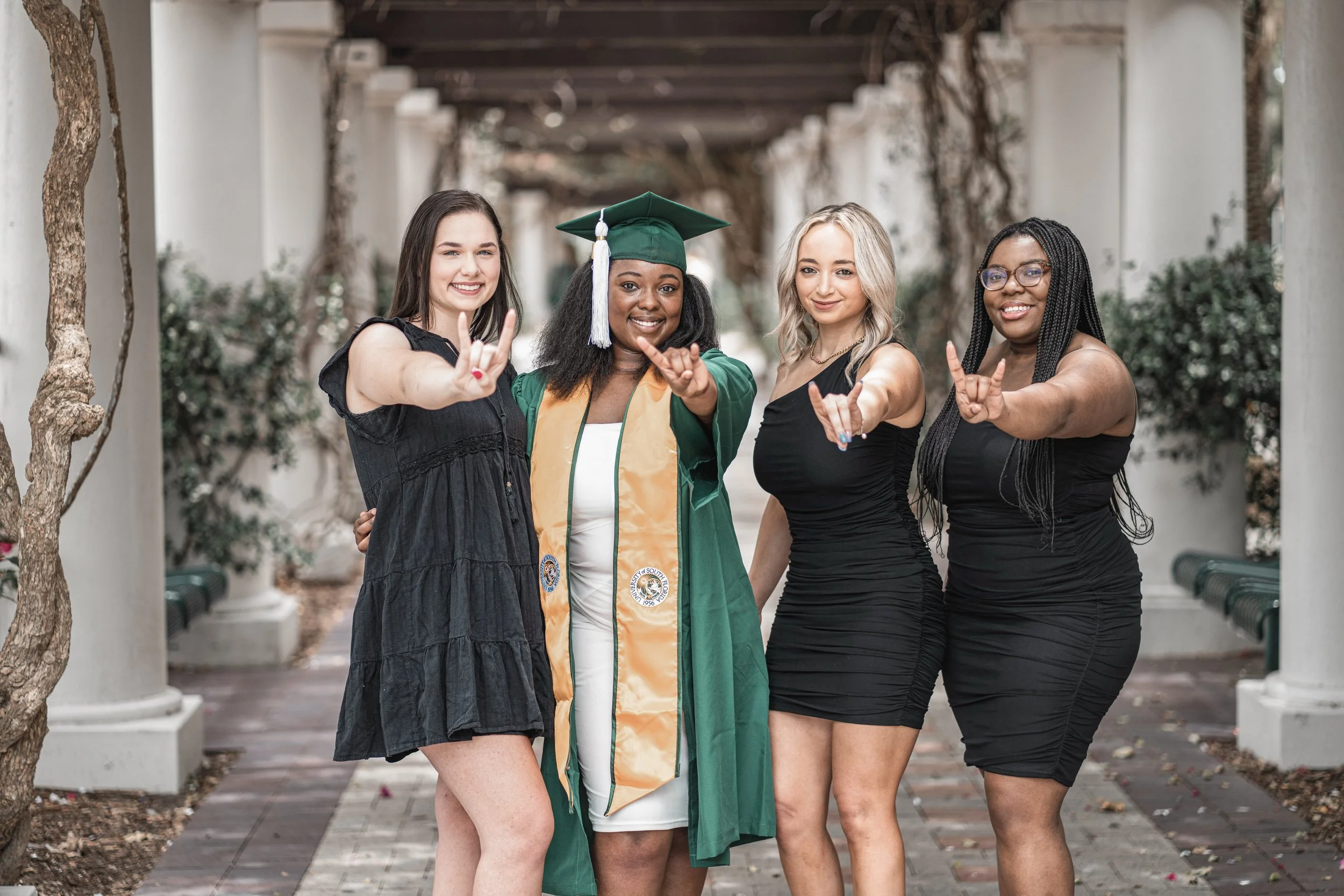 Four young women standing in an outdoor walkway, with one wearing a graduation cap and gown, all smiling and making rock and roll hand gestures.