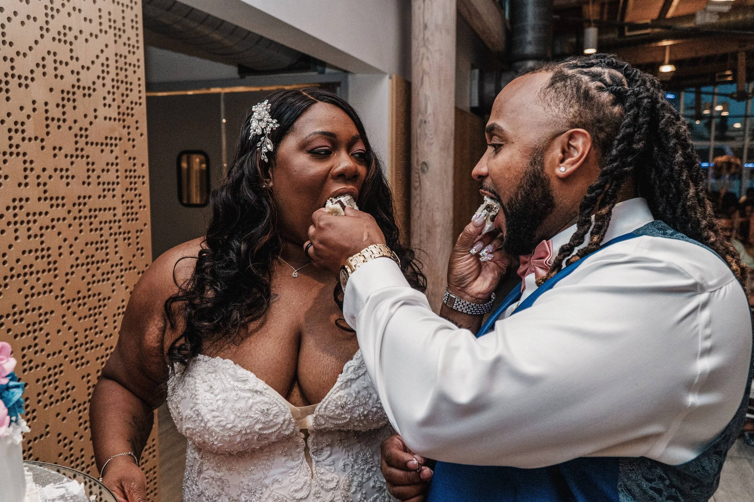 A bride and groom, both with dark skin, sharing a moment during their wedding celebration. The bride is wearing a white lace wedding dress with curly dark hair adorned with a decorative hairpiece, and the groom is dressed in a white tuxedo shirt with