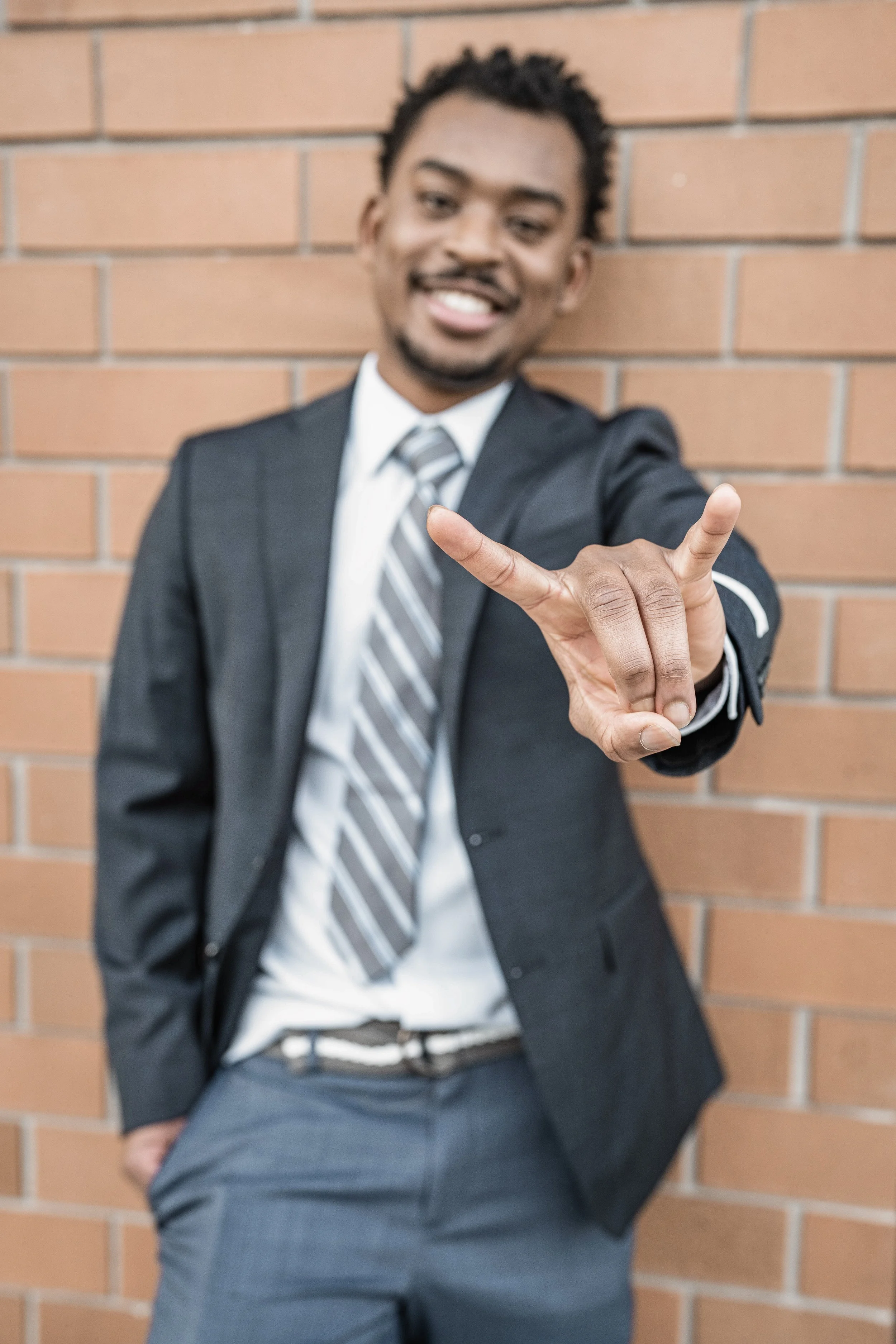 A man in a suit smiling and making a rock and roll hand gesture with his right hand, standing against a brick wall.