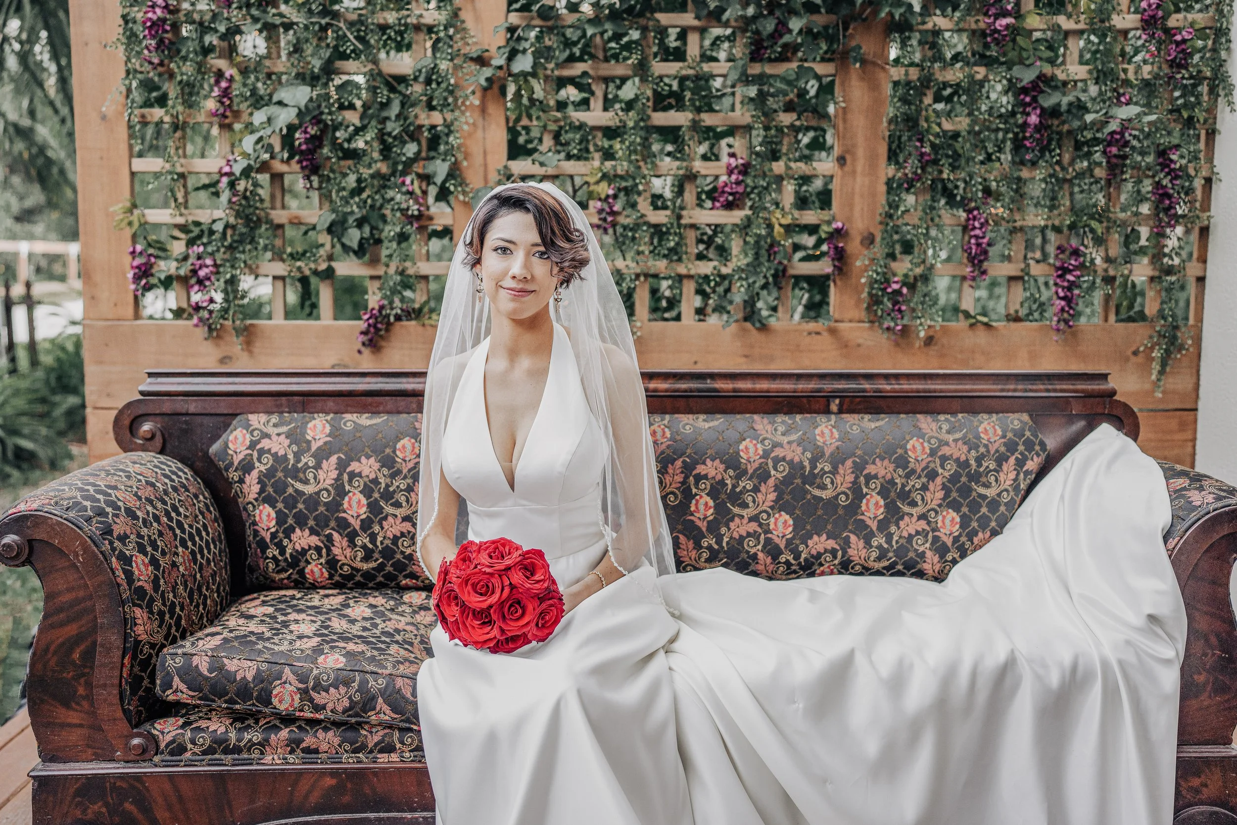 A woman in a wedding dress sitting on a vintage floral sofa, holding a bouquet of red roses, with a wooden lattice and greenery in the background.