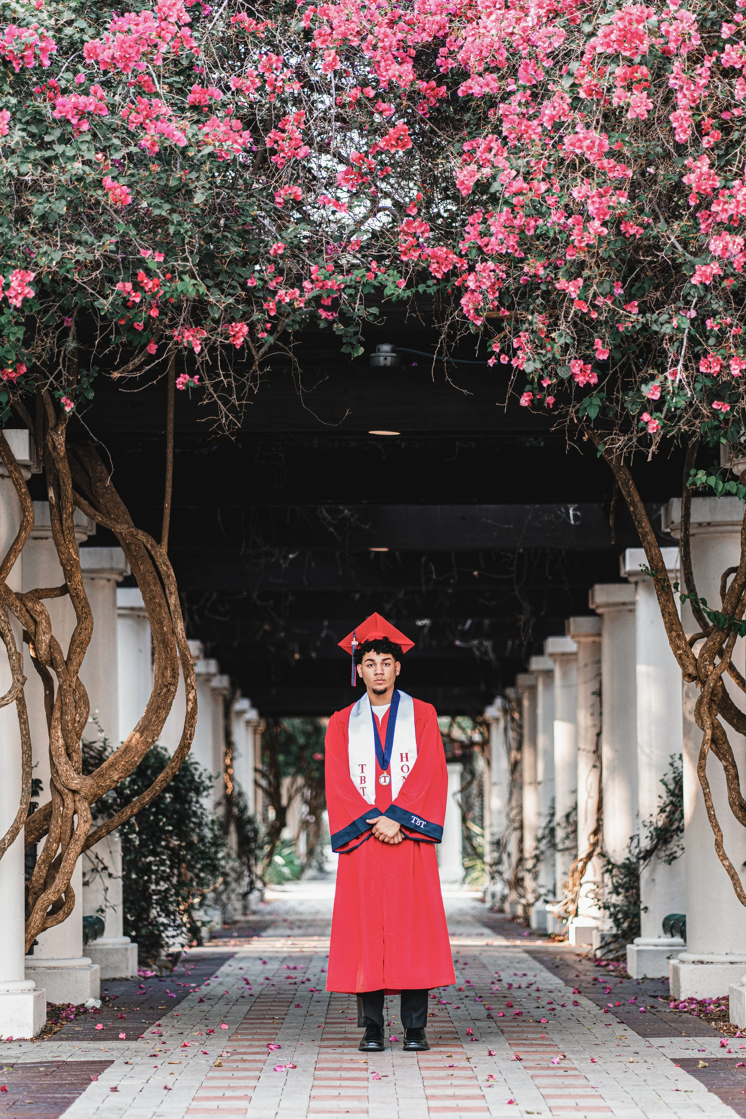 A young man in a red graduation gown and cap stands under a floral archway with pink flowers, on a pathway lined with flower petals.