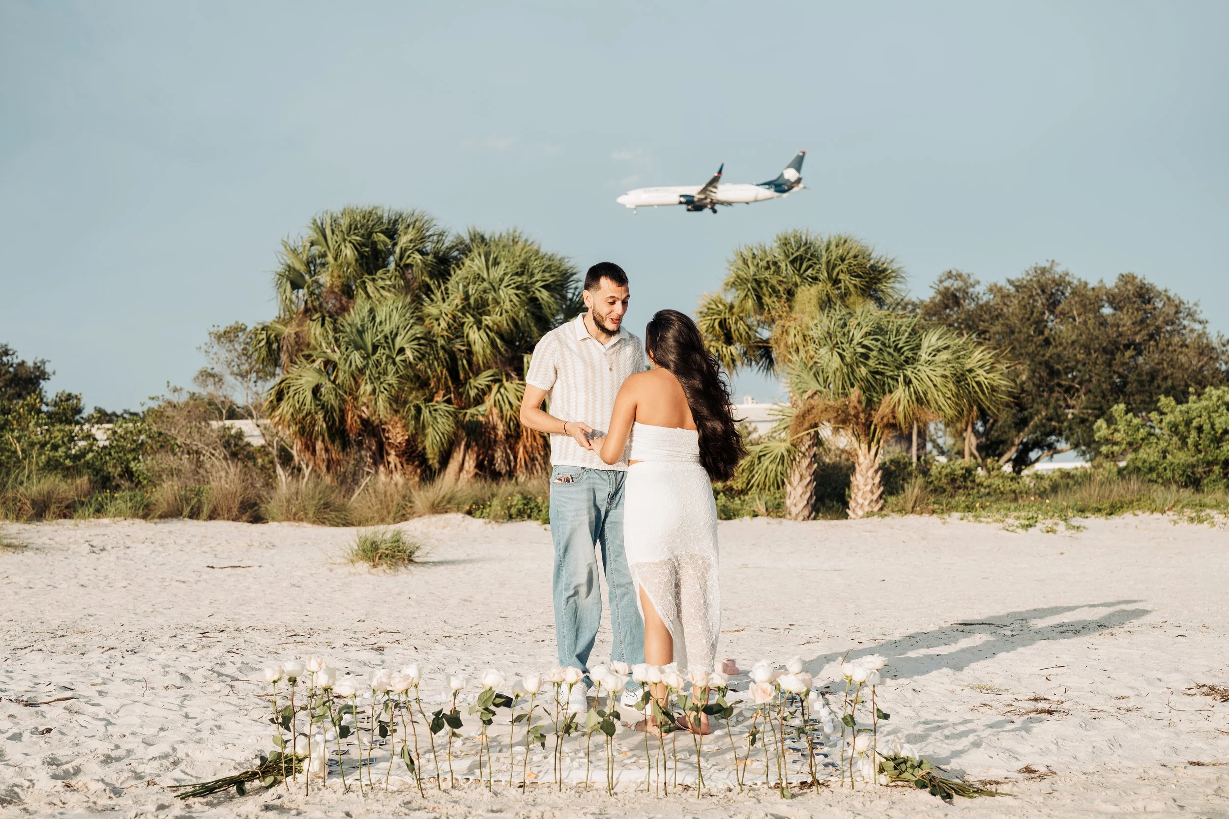 A couple standing on a sandy beach with palm trees, with an airplane flying overhead in the sky.