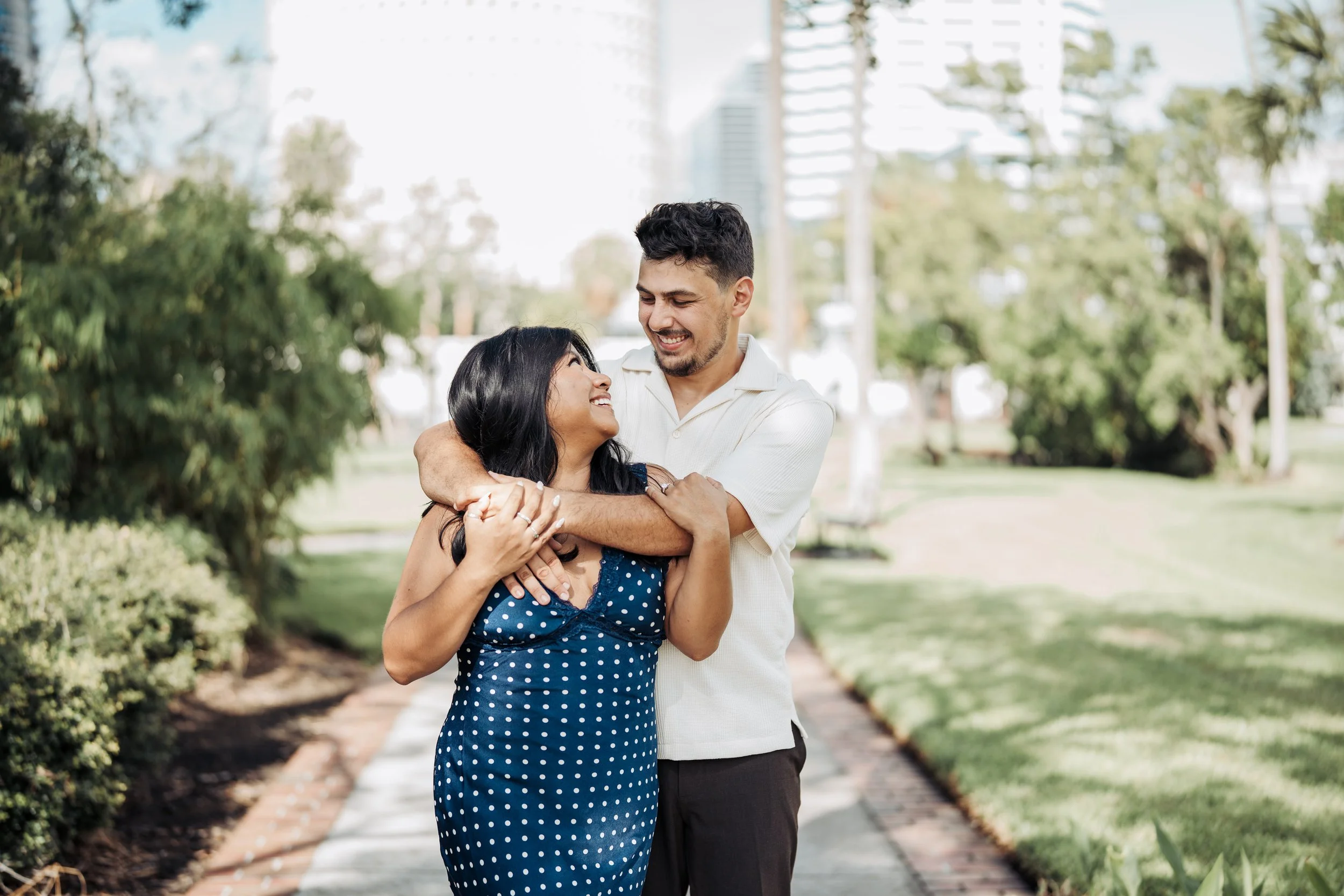 A smiling woman in a blue polka dot dress and a smiling man in a white shirt embrace outdoors in a park with trees, grass, and city buildings in the background.