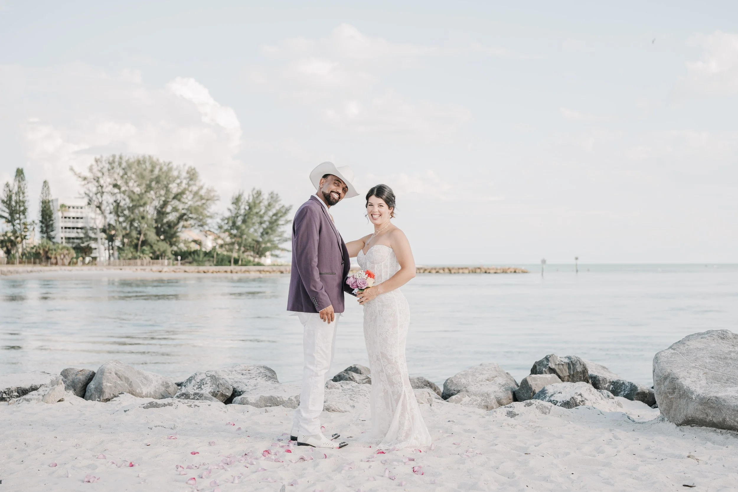 A couple in wedding attire standing on rocks by the beach with water, trees, and buildings in the background.
