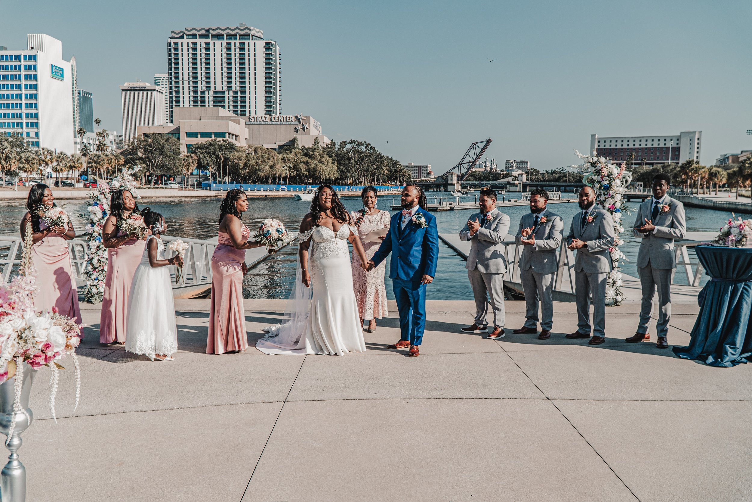 A wedding ceremony taking place outdoors by the water with city high-rise buildings in the background. The bride and groom are holding hands, surrounded by bridesmaids and groomsmen dressed in formal attire. Floral decorations are visible.