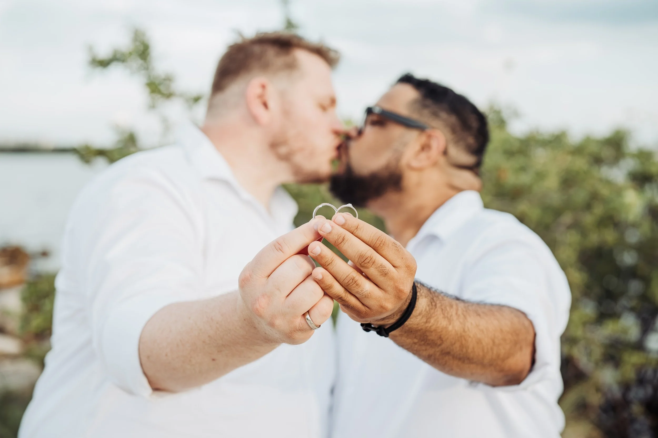 Two men are holding a pair of wedding rings and kissing each other outdoors, with a background of trees and sky.