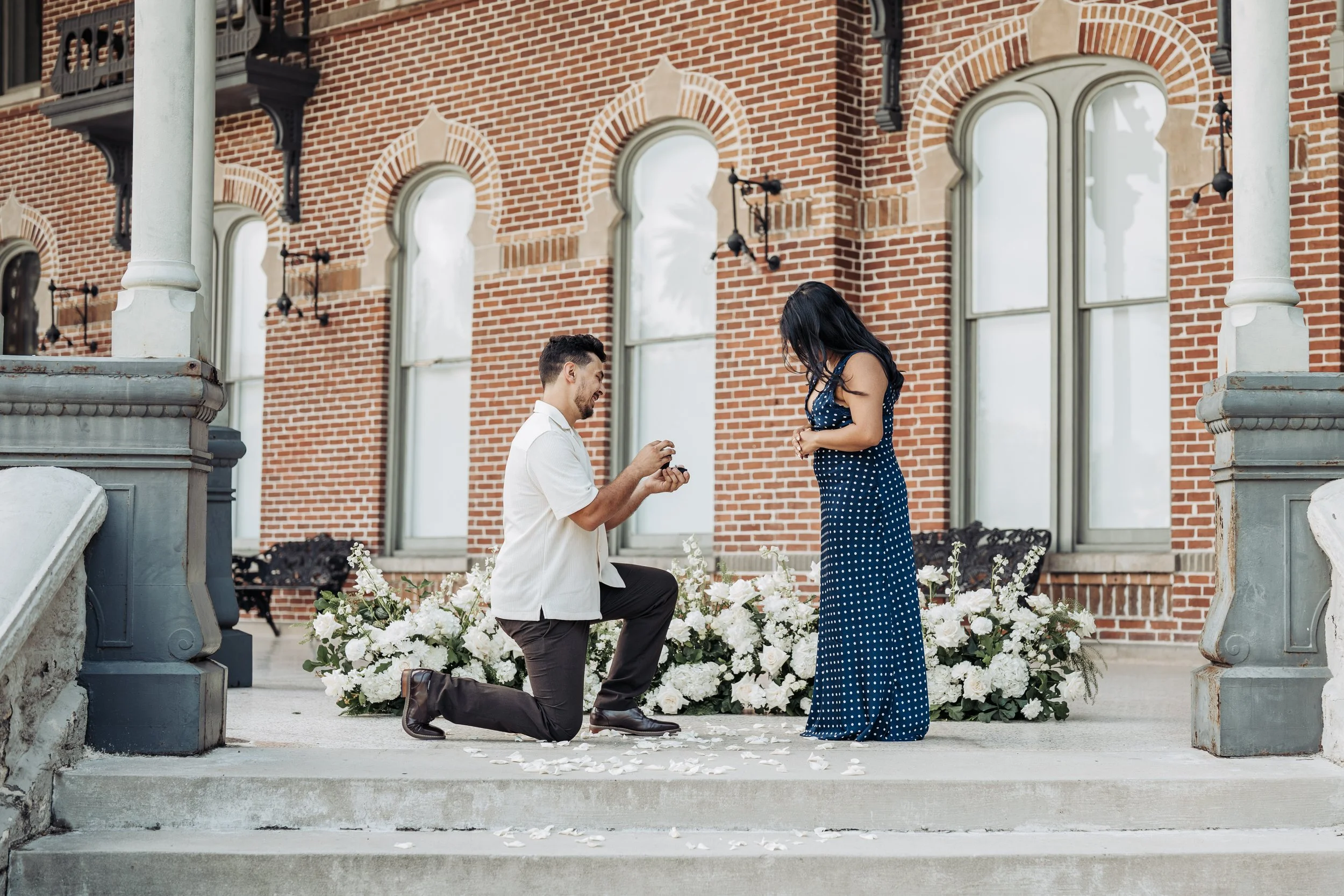 A man proposing marriage to a woman on the steps of a brick building, with floral decorations around them and a person on one knee offering a ring.