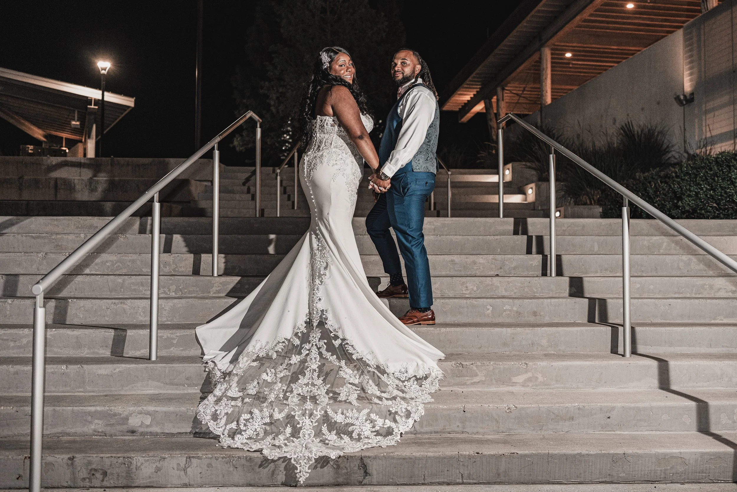 A bride and groom holding hands and standing on stairs outdoors at night, smiling and looking at each other. The bride is wearing a white lace wedding gown with a long train, and the groom is in a dark blue vest, white shirt, and blue pants. There ar
