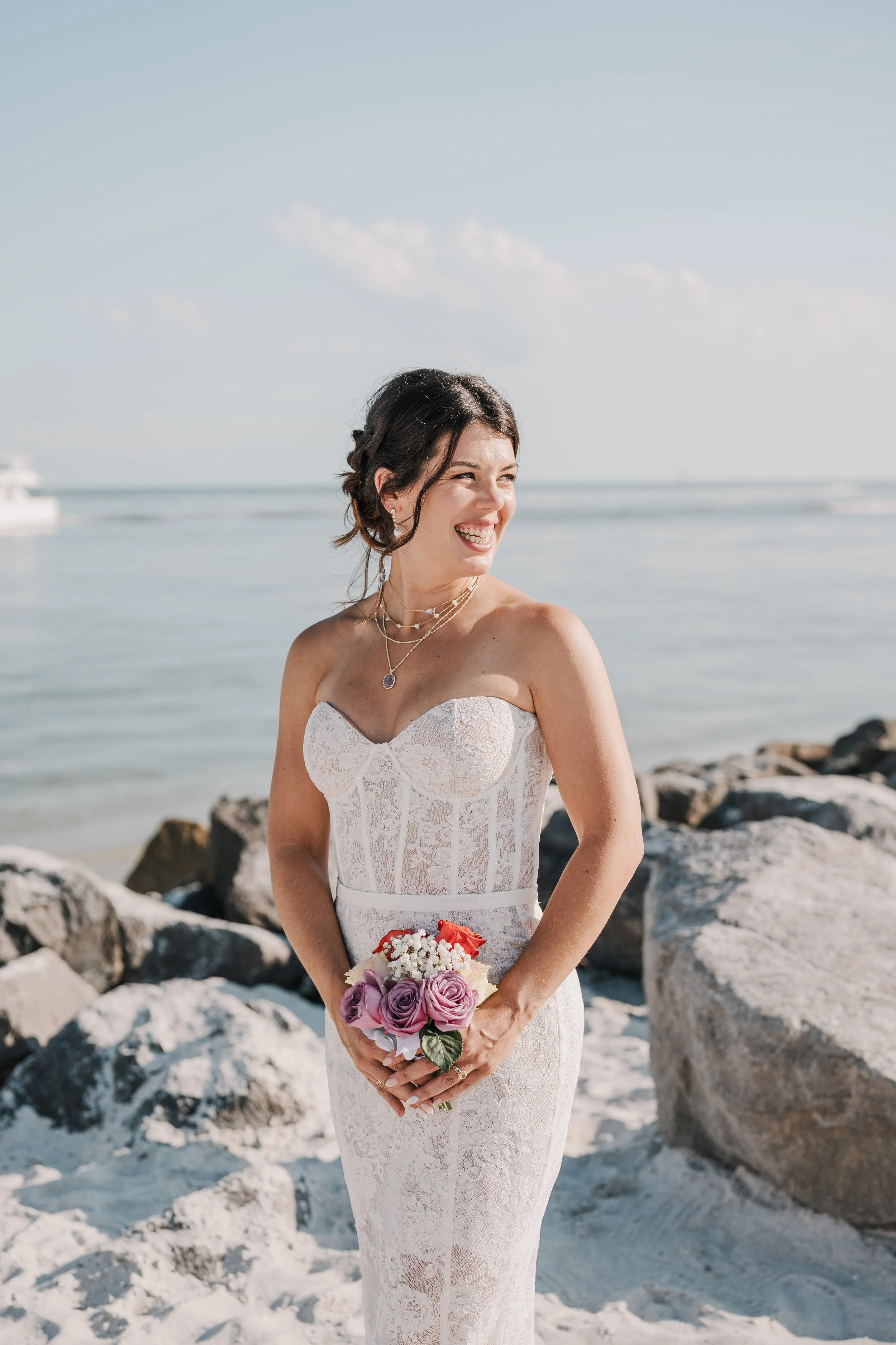 A bride in a white lace wedding dress holding a bouquet of pink and purple roses, standing on a rocky beach with ocean water and a boat in the background.
