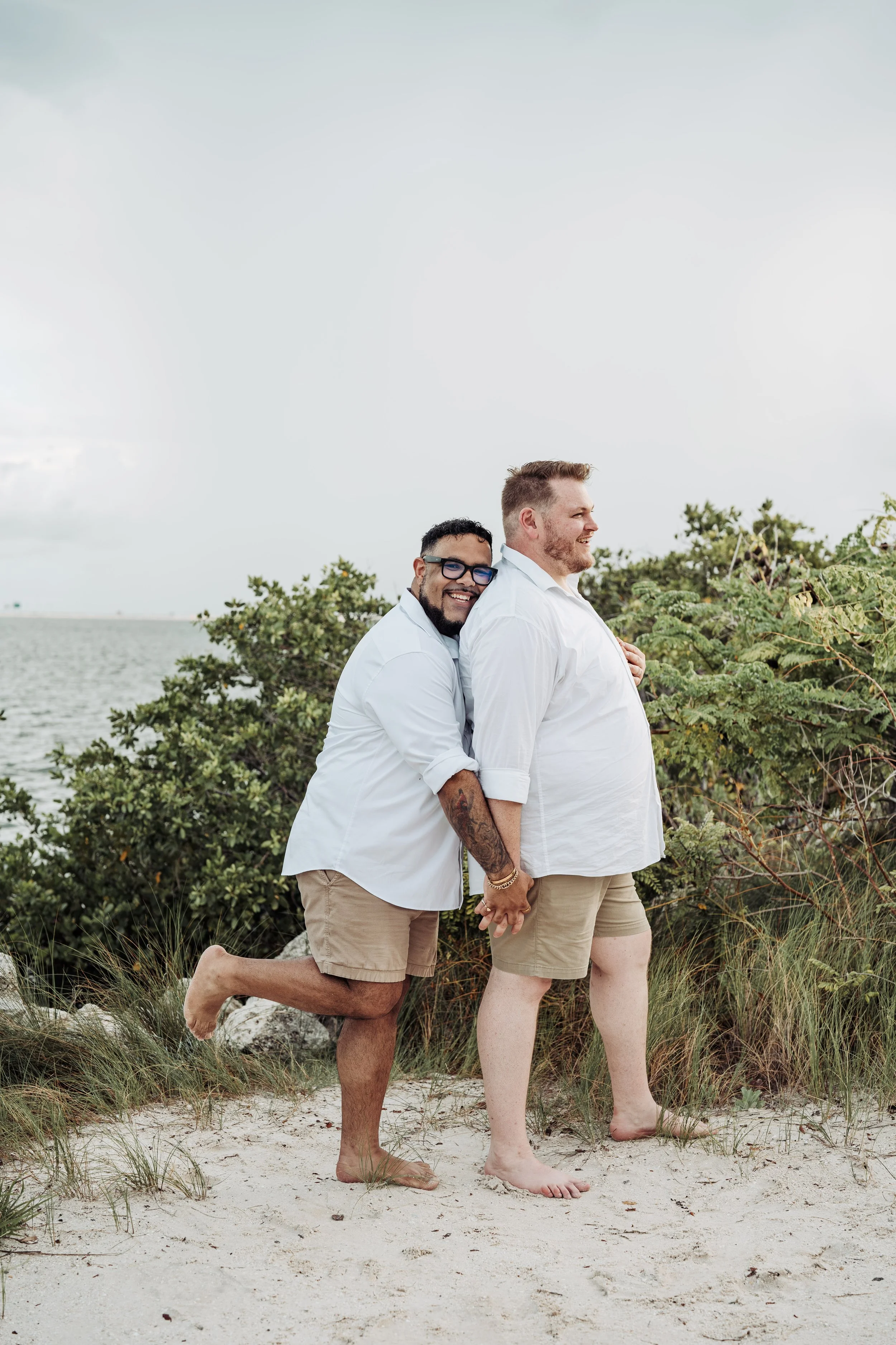 Two men holding hands, hugging, and smiling on a beach with green bushes, sand, and the ocean in the background. One man is kneeling while the other stands, both wearing white shirts and beige shorts.
