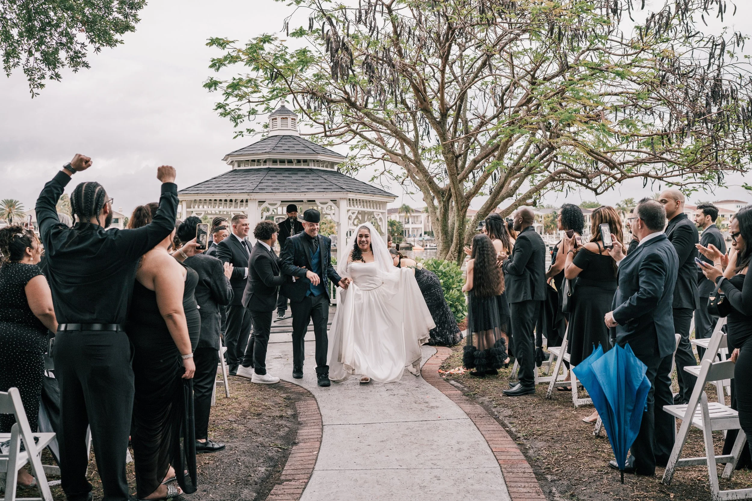 A bride and groom holding hands walking down the aisle at an outdoor wedding ceremony, surrounded by guests. The scene is set near a gazebo with trees and a cloudy sky in the background.