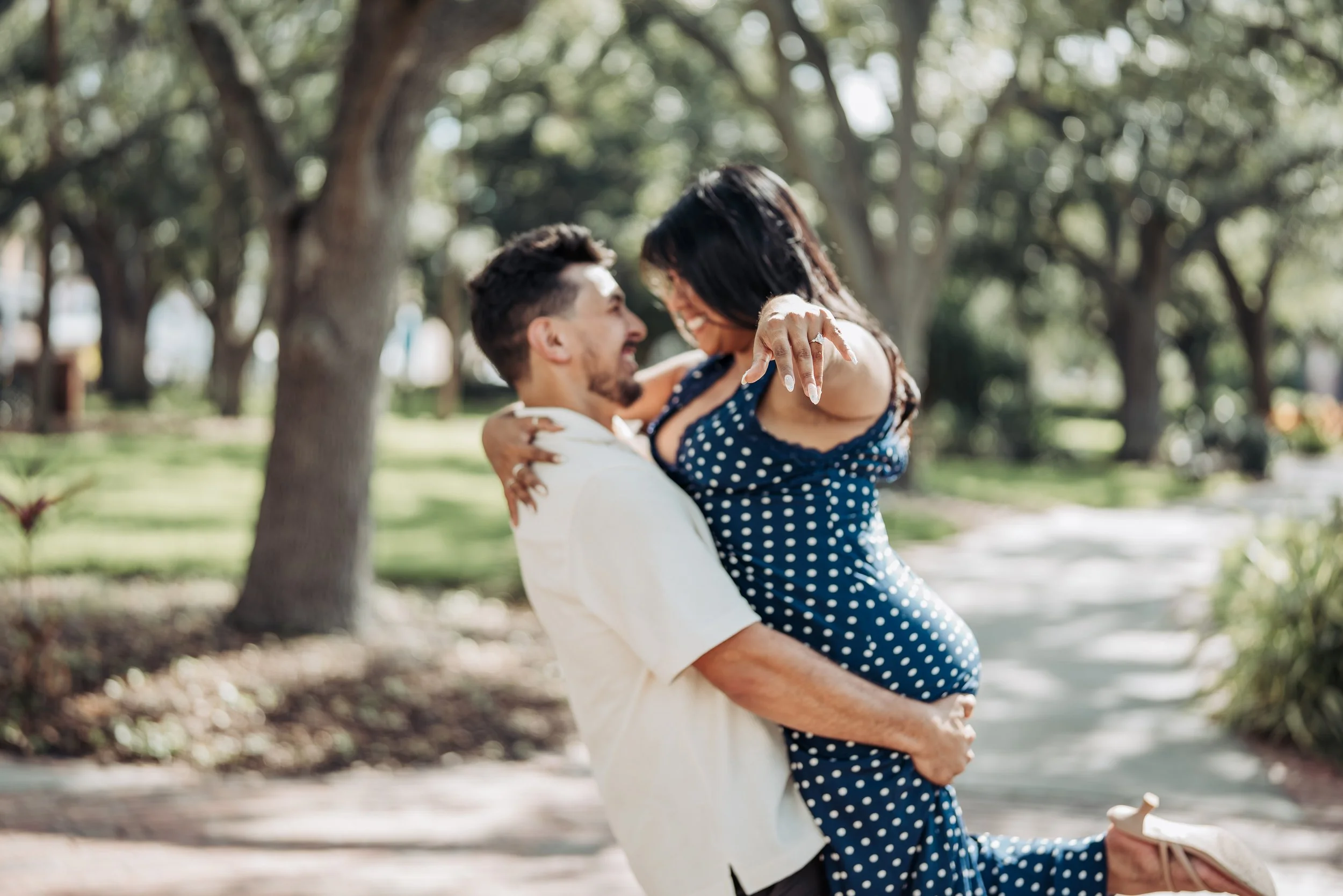 A happy couple outdoors in a park, with the man lifting the woman in his arms, both smiling and engaged in a joyful moment.
