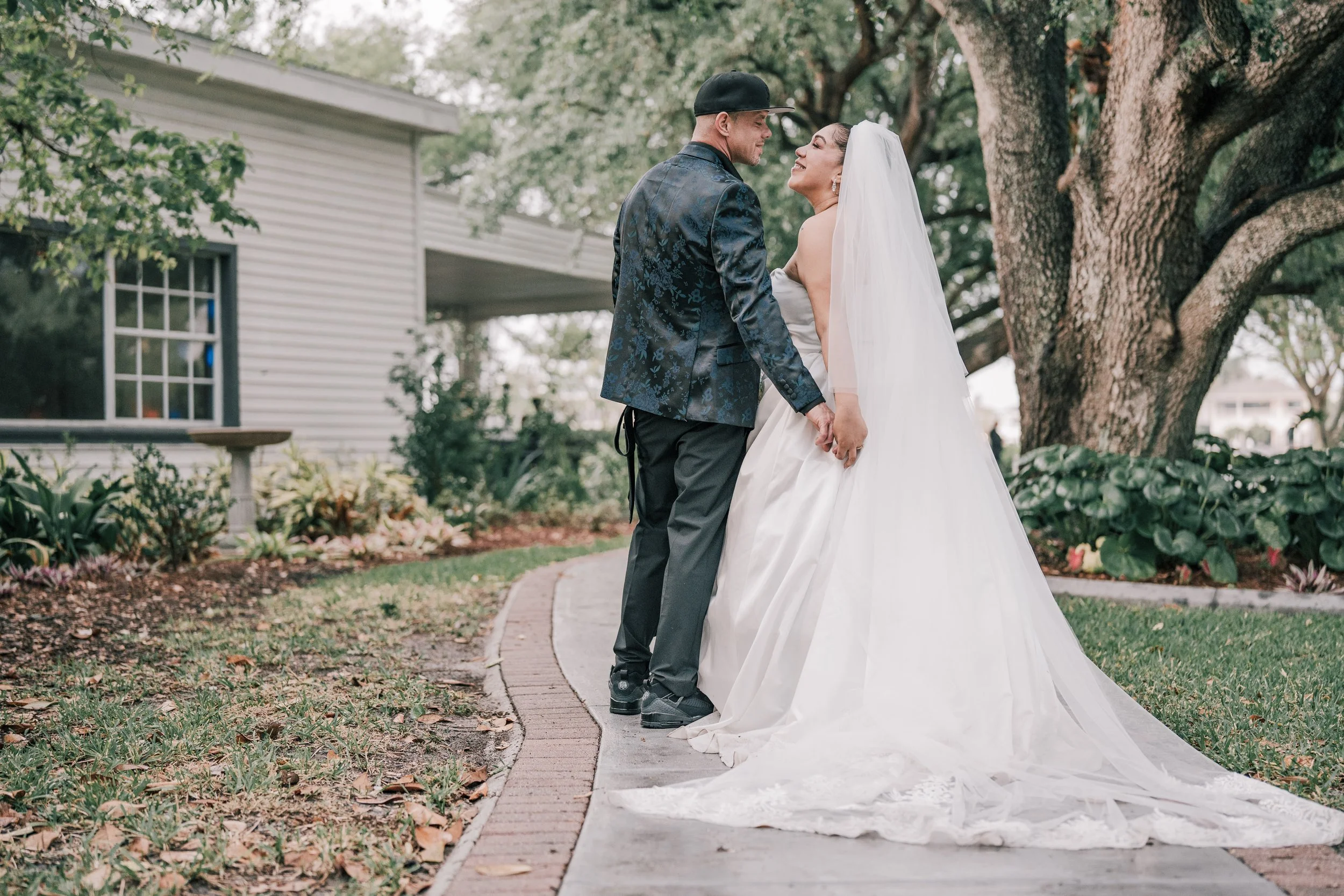 A bride and groom standing close together outdoors, smiling at each other. The bride wears a white wedding dress and veil, and the groom wears a dark blue patterned blazer, gray pants, and a black cap. They are holding hands beneath a large tree, wit