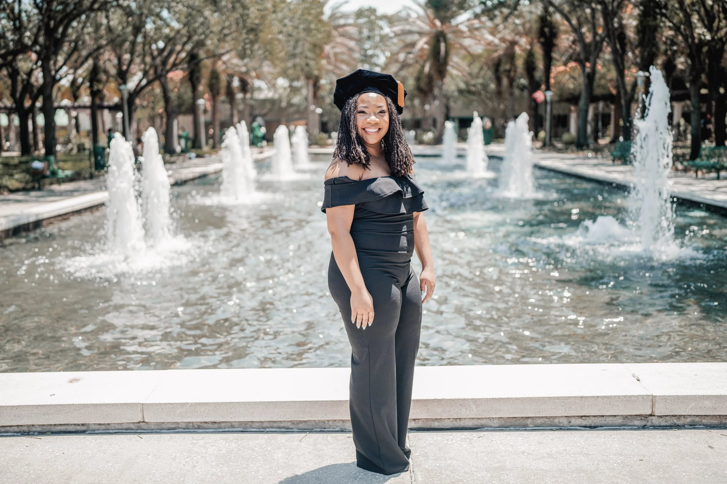 A woman in a black off-shoulder dress and graduation cap standing in front of a fountain in a park with trees in the background.