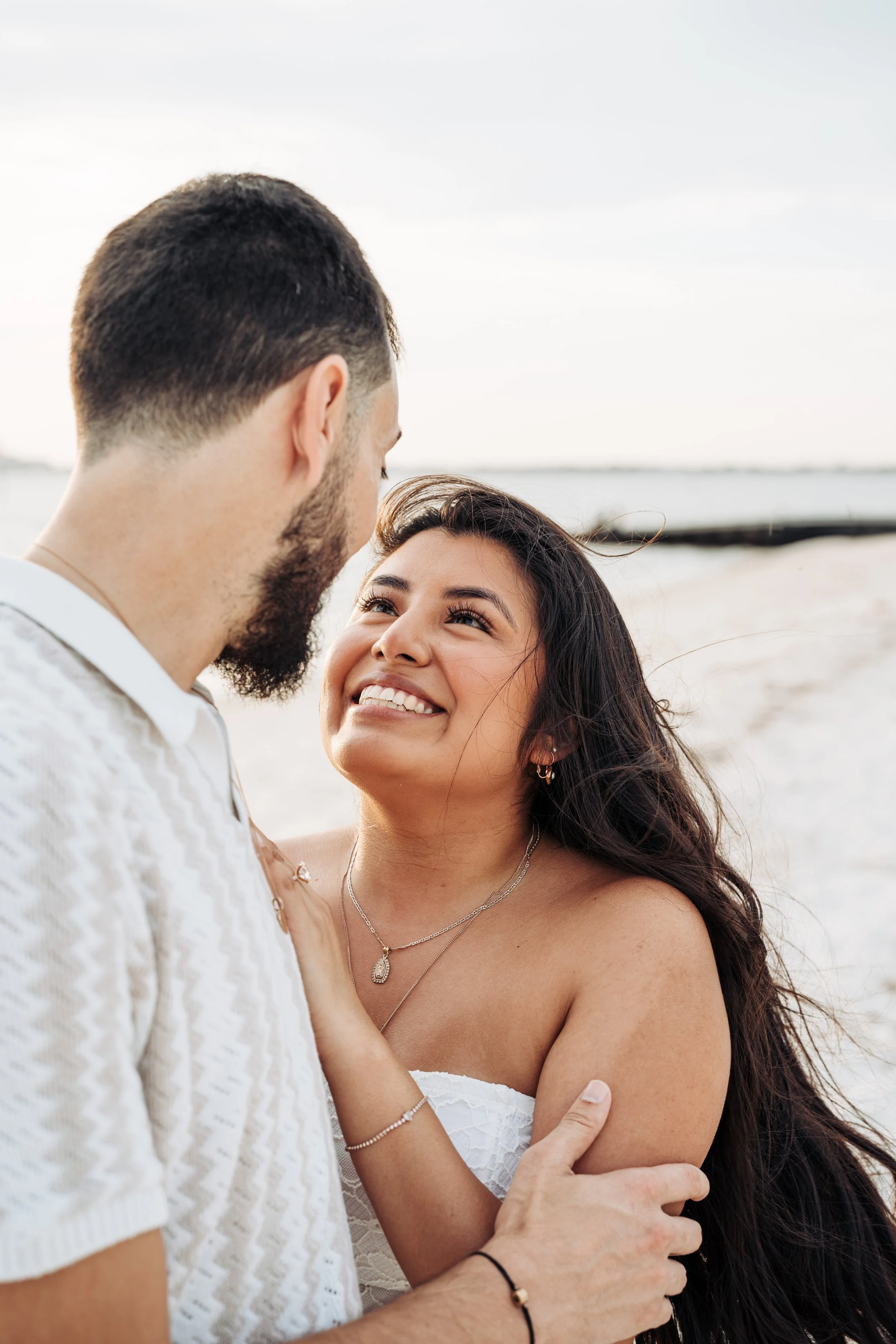 A smiling woman looking up at a man on a beach, both embracing each other closely.