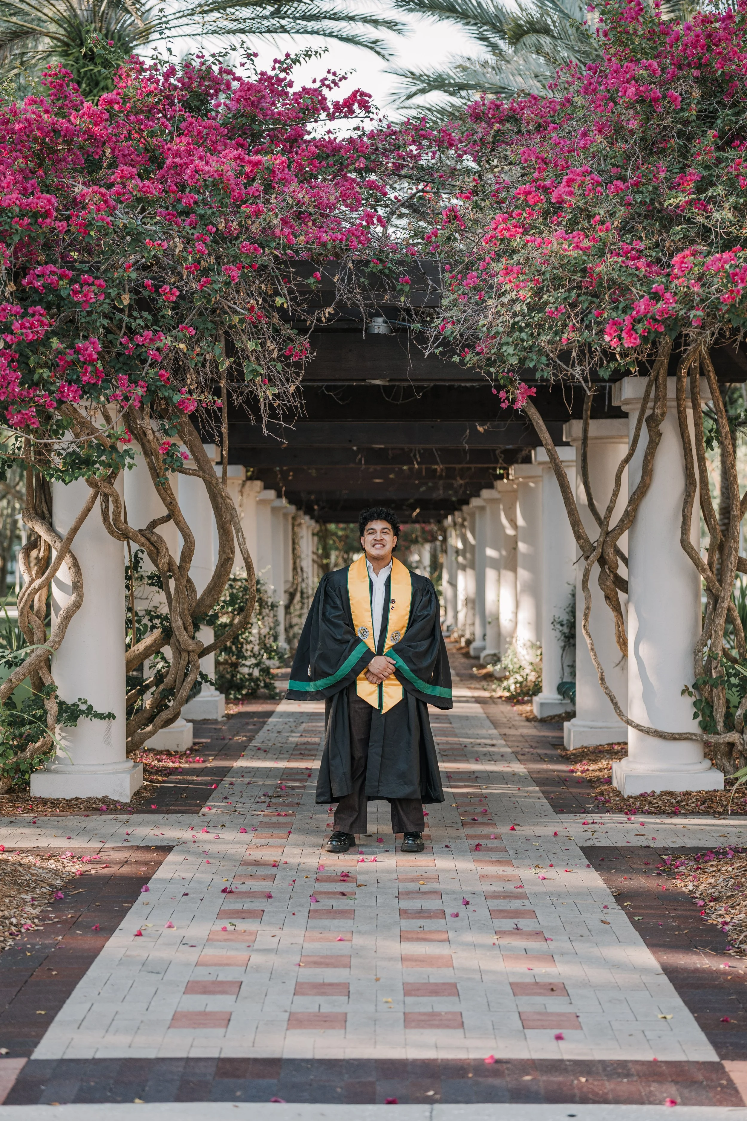 A smiling man in a graduation gown and cap standing in a garden pathway, amidst pink flowering bushes and white columns.