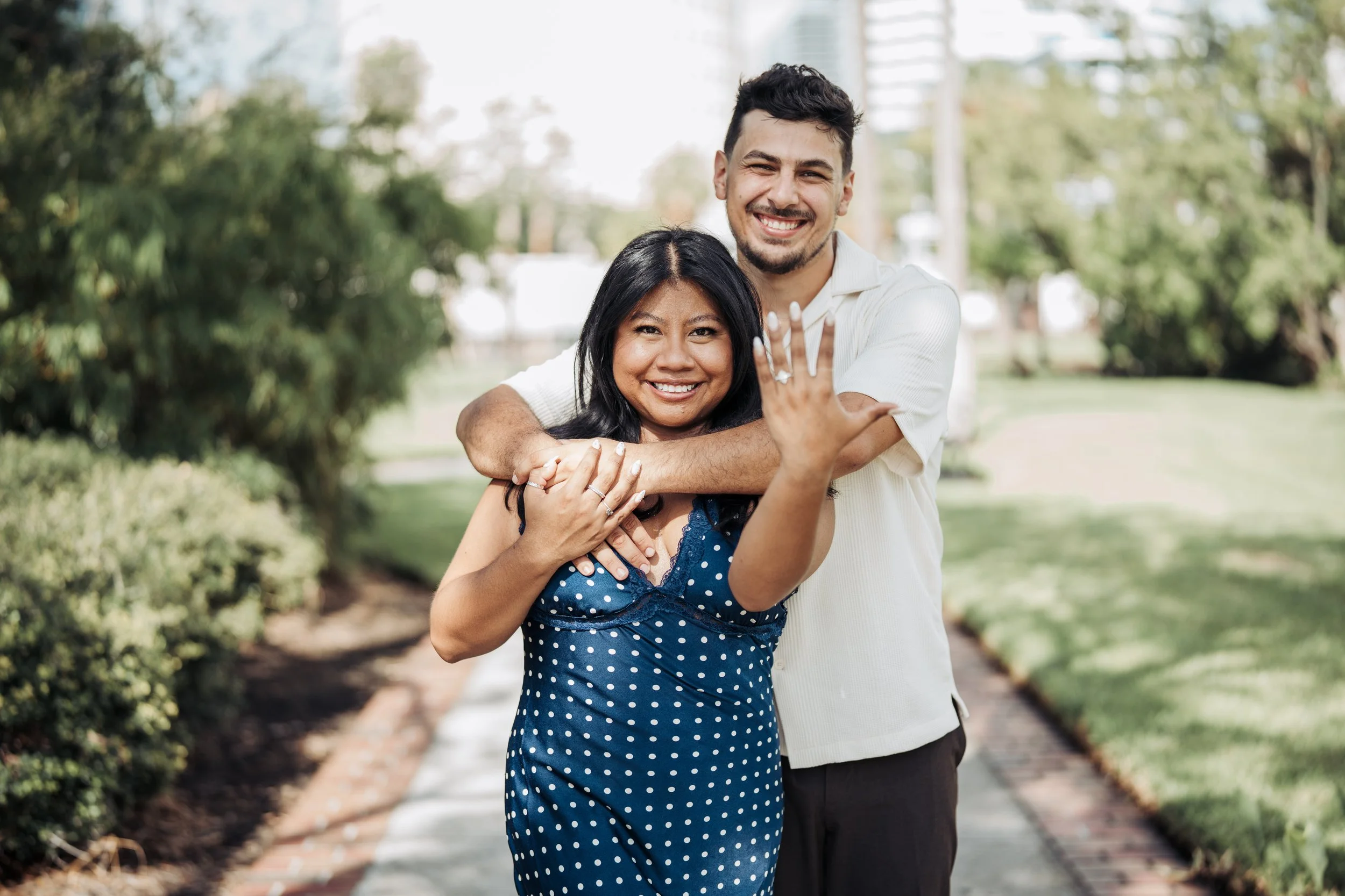 A smiling couple standing outdoors in a park, with the woman showing off her engagement ring and the man hugging her from behind.